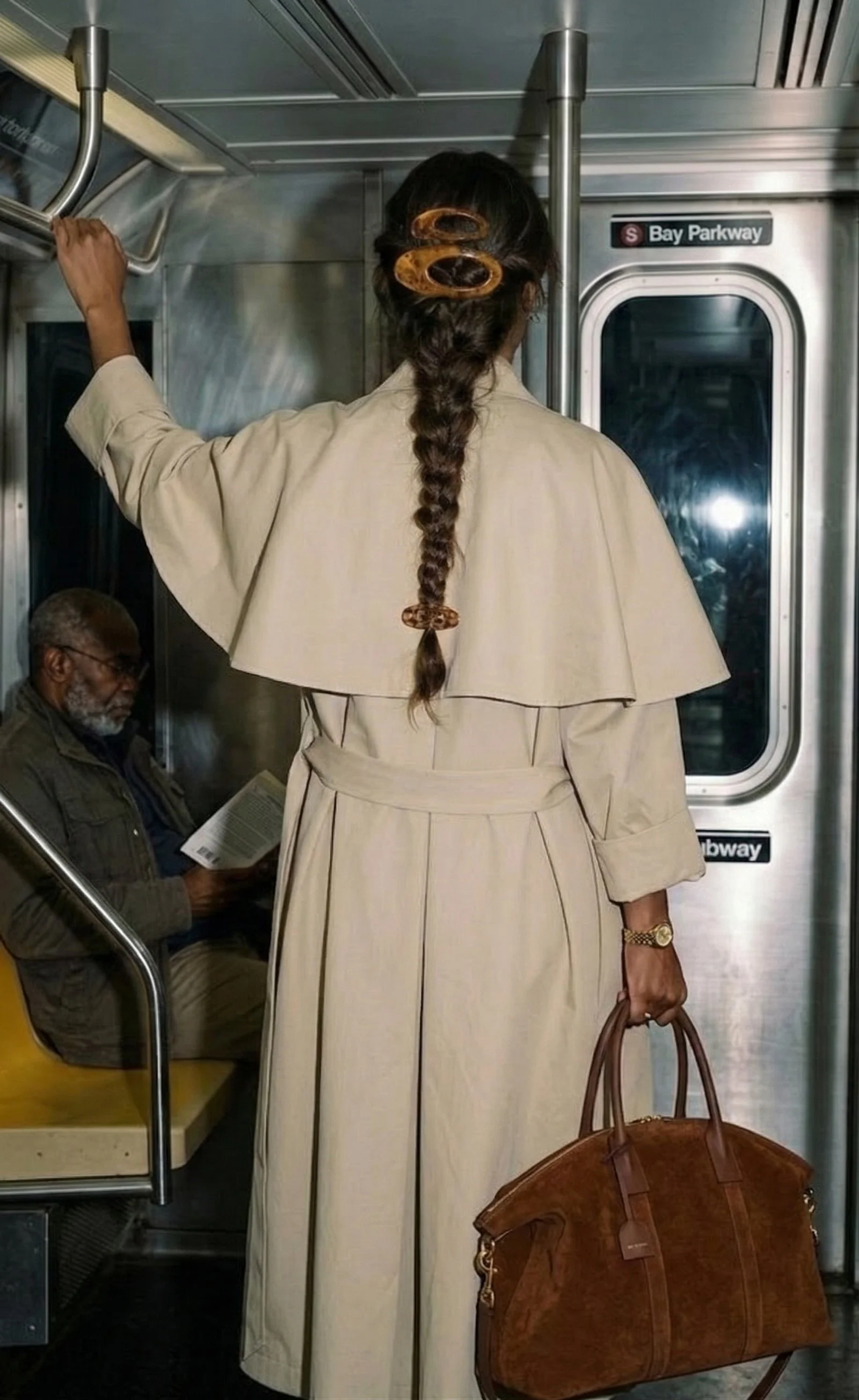 A woman in a beige trench coat standing in a subway car, holding onto a handrail with one hand and carrying a large brown handbag in the other, with an older man reading a book seated in the background.