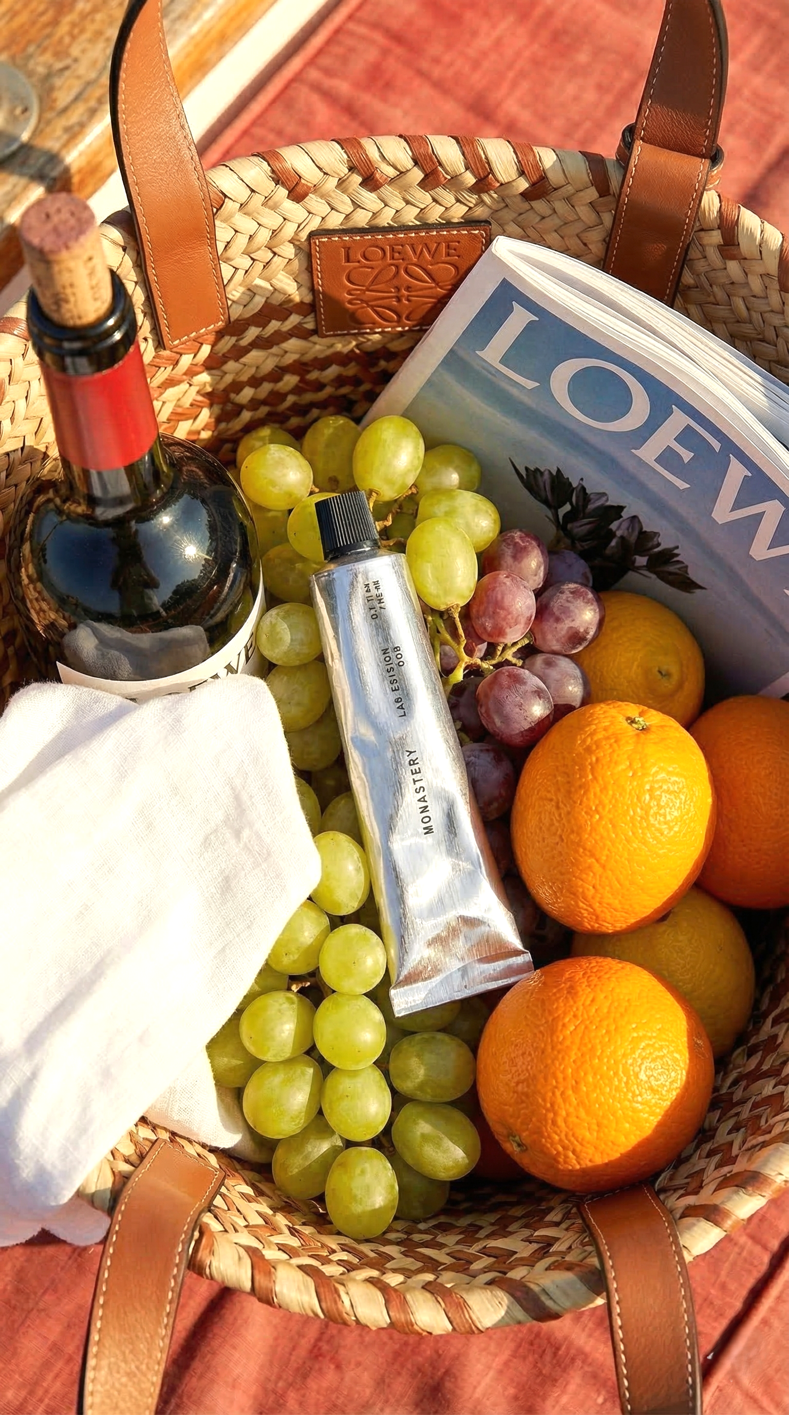 A woven basket containing green and red grapes, oranges, a bottle of wine, a tube of skincare product, LOEWE magazine, and a white cloth.