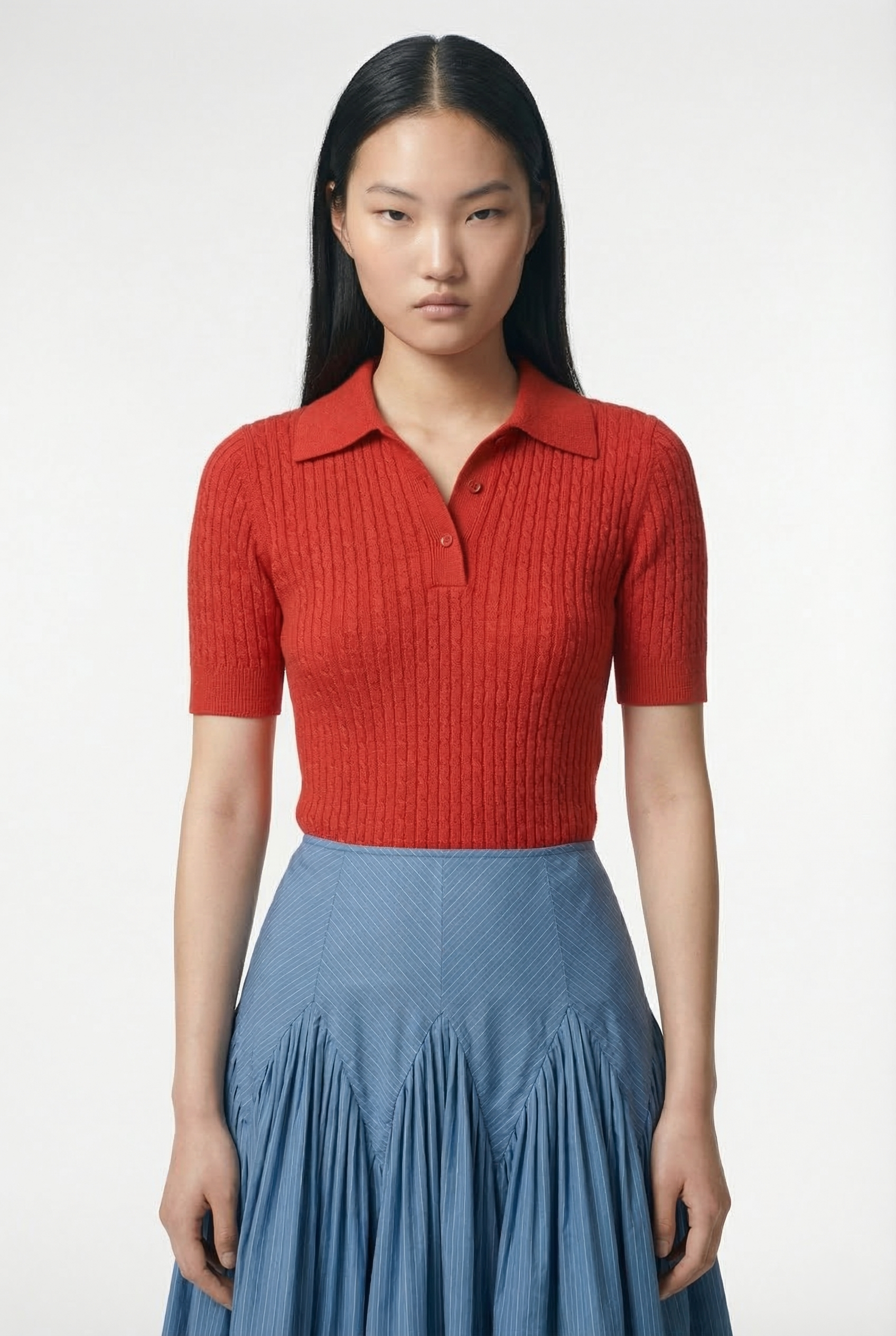 Young woman with long black hair wearing a fitted red short-sleeve polo shirt and a blue pleated skirt.