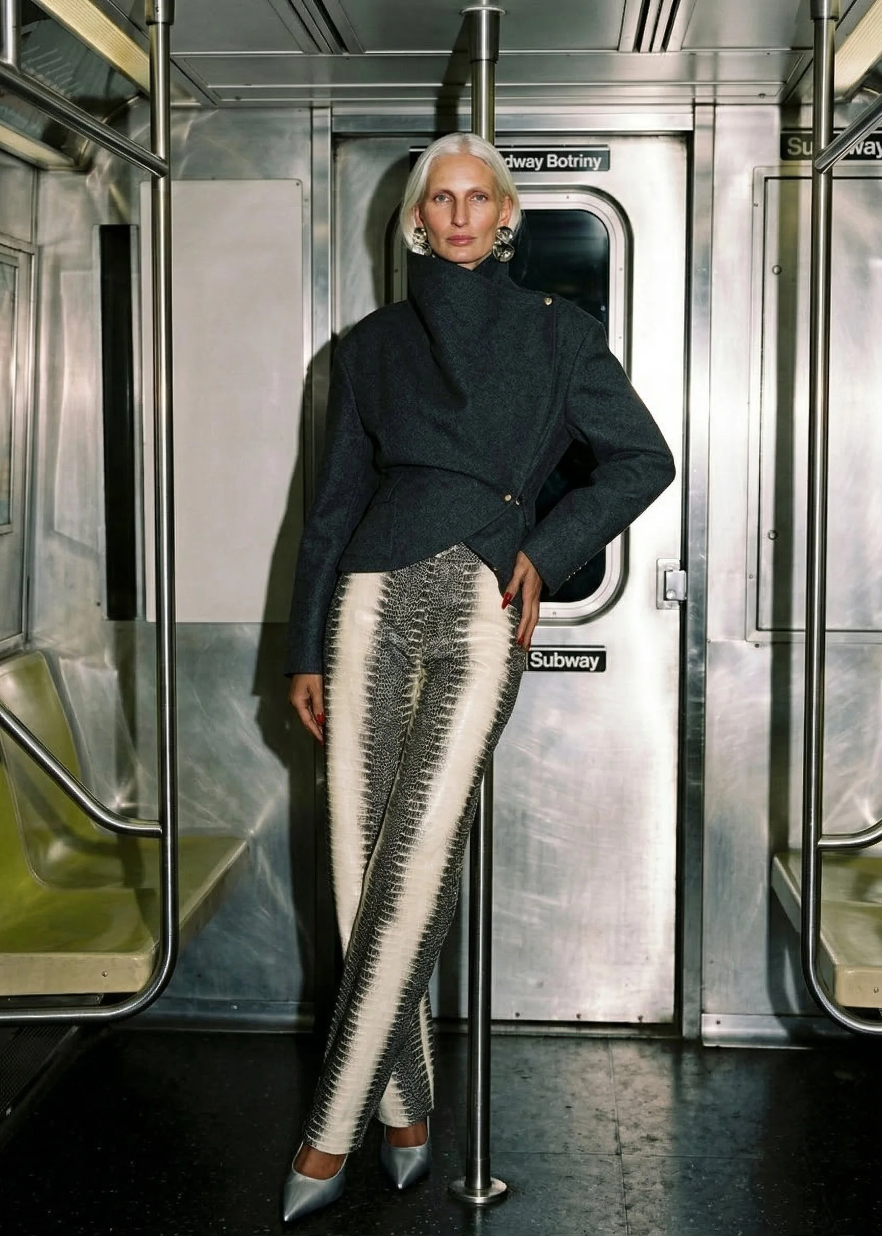 Woman with silver hair and large earrings standing inside a subway train, wearing a dark coat, patterned pants, and silver high heels.