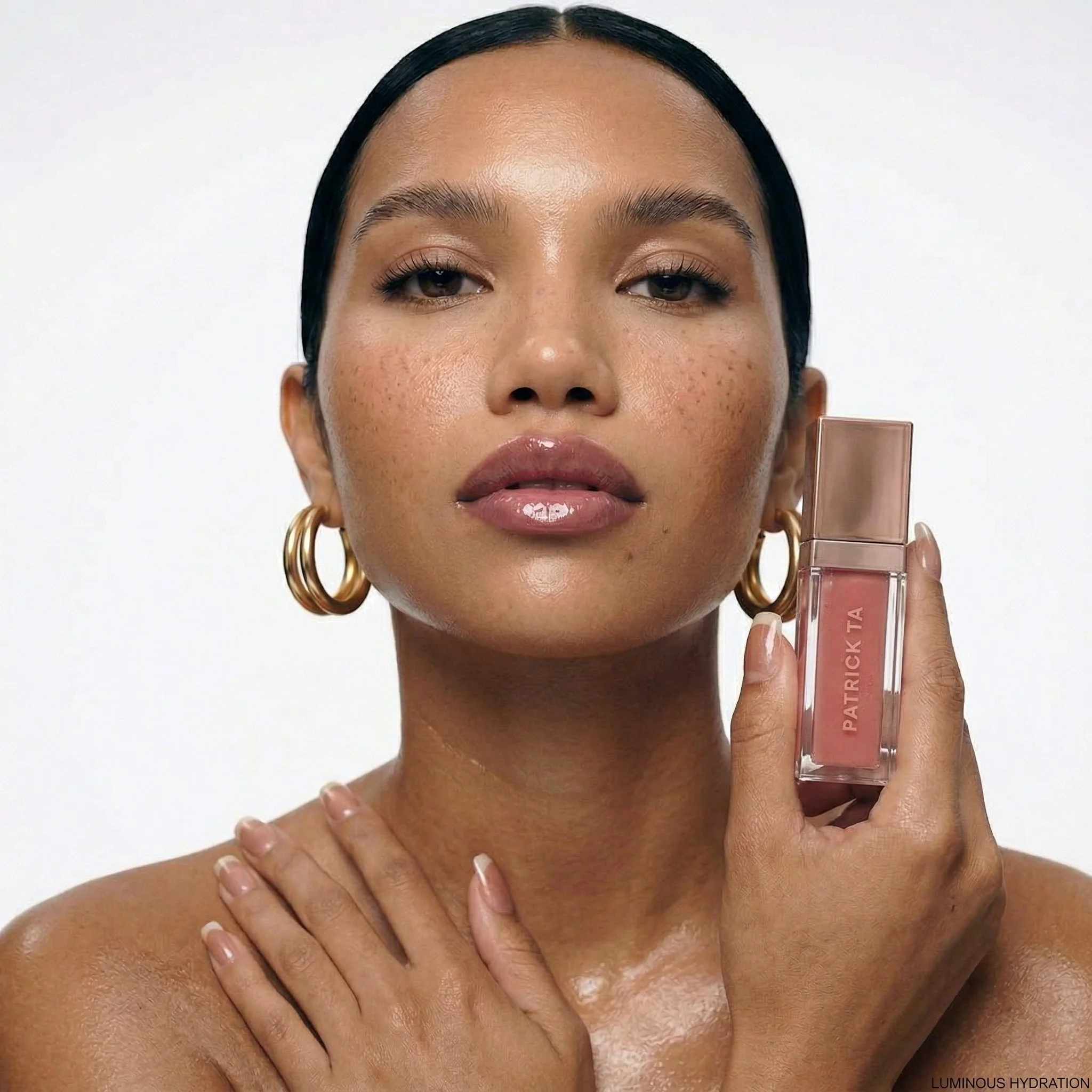 Close-up of a woman with glossy lips and gold hoop earrings holding a bottle of Patrick Ta blush, looking directly at the camera with a neutral background.