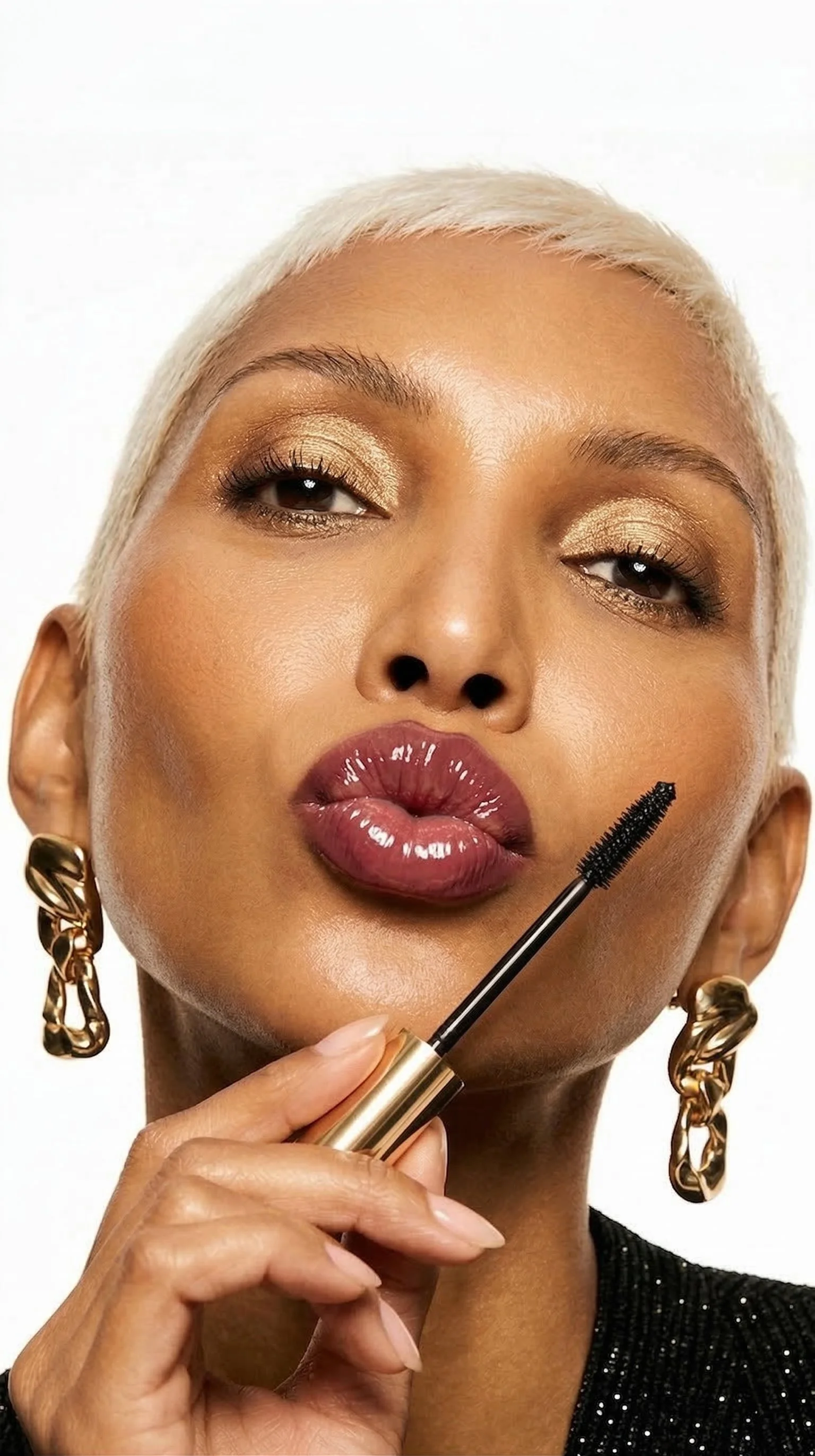 Close-up of a woman with platinum blonde hair applying mascara to her eyelashes, holding the mascara wand near her face, wearing gold earrings and makeup.