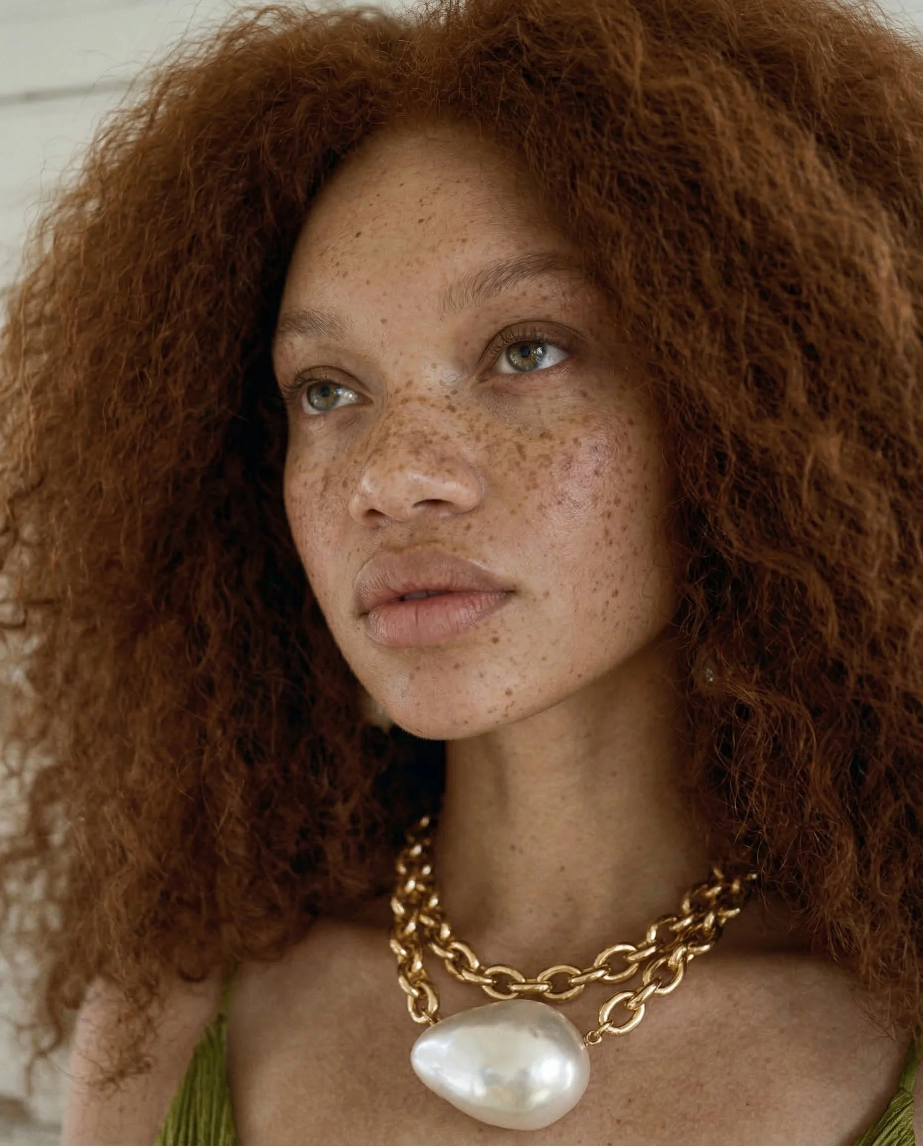 Close-up of a woman with curly red hair, freckles, and blue eyes, wearing a chunky gold chain necklace with a large pearl pendant and a green top.