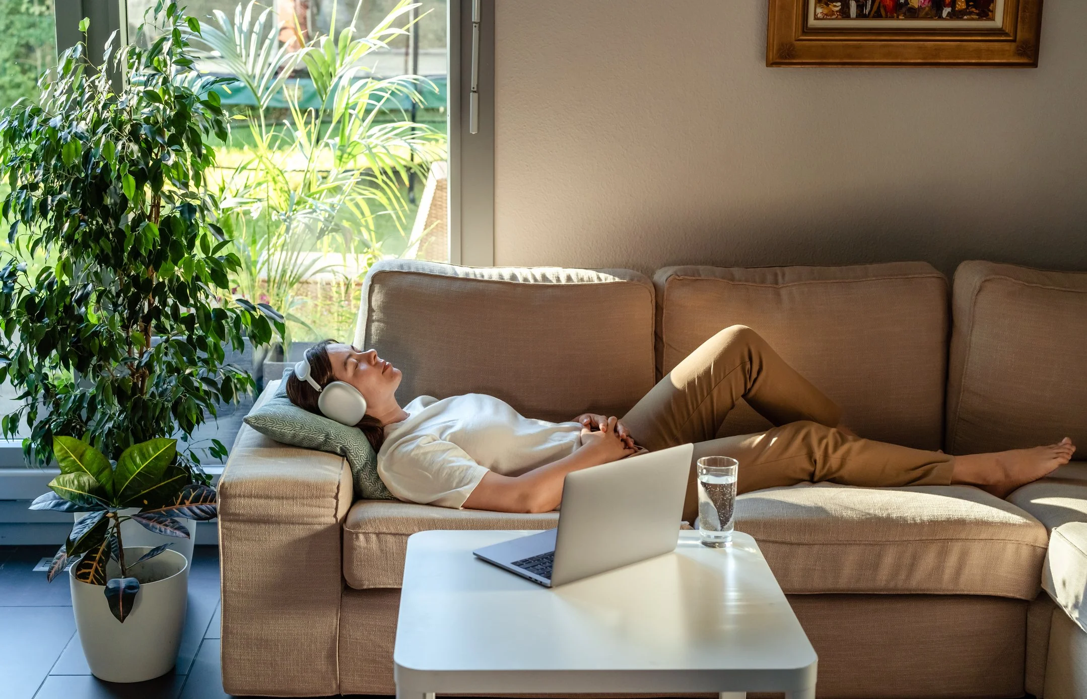 A woman laying on a beige sofa, resting with her eyes closed, wearing headphones, with a laptop and a glass of water on a small white table nearby, in a room with large windows and green plants outside.