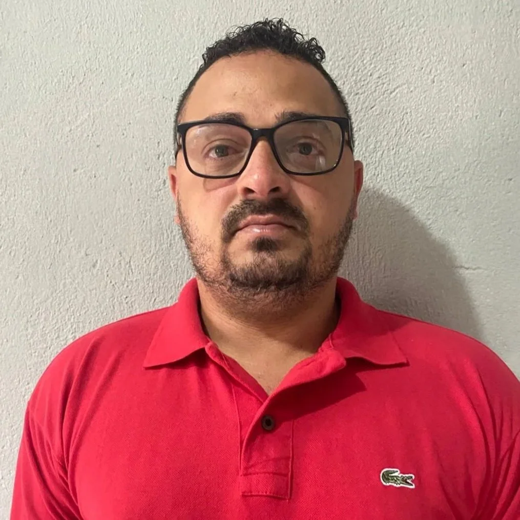 Homem com barba, cabelo curto, usando óculos escuros, camiseta vermelha da Rip Curl, e crachá no pescoço, posando em frente a uma parede branca com uma régua de altura ao lado.