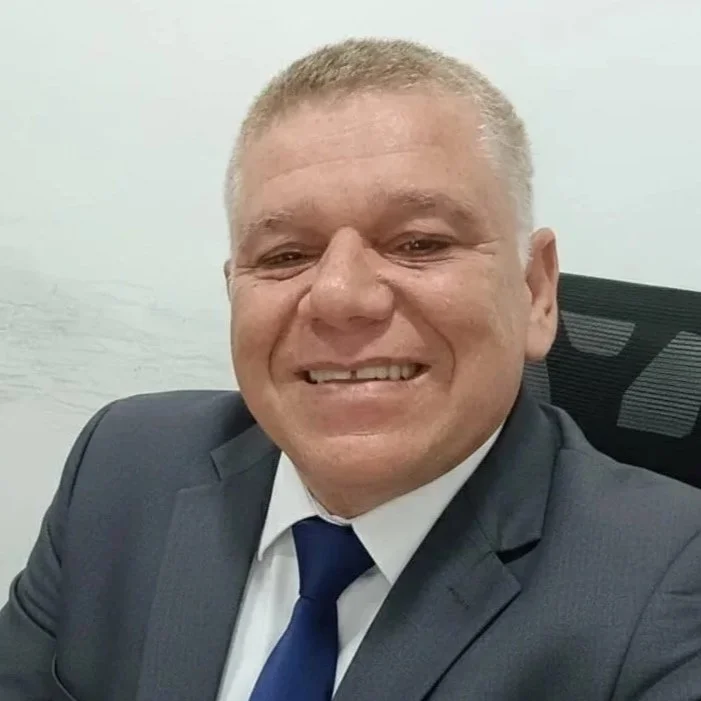 Homem com cabelo curto grisalho, rosto sorridente, usando camisa azul clara, fundo de parede branca.