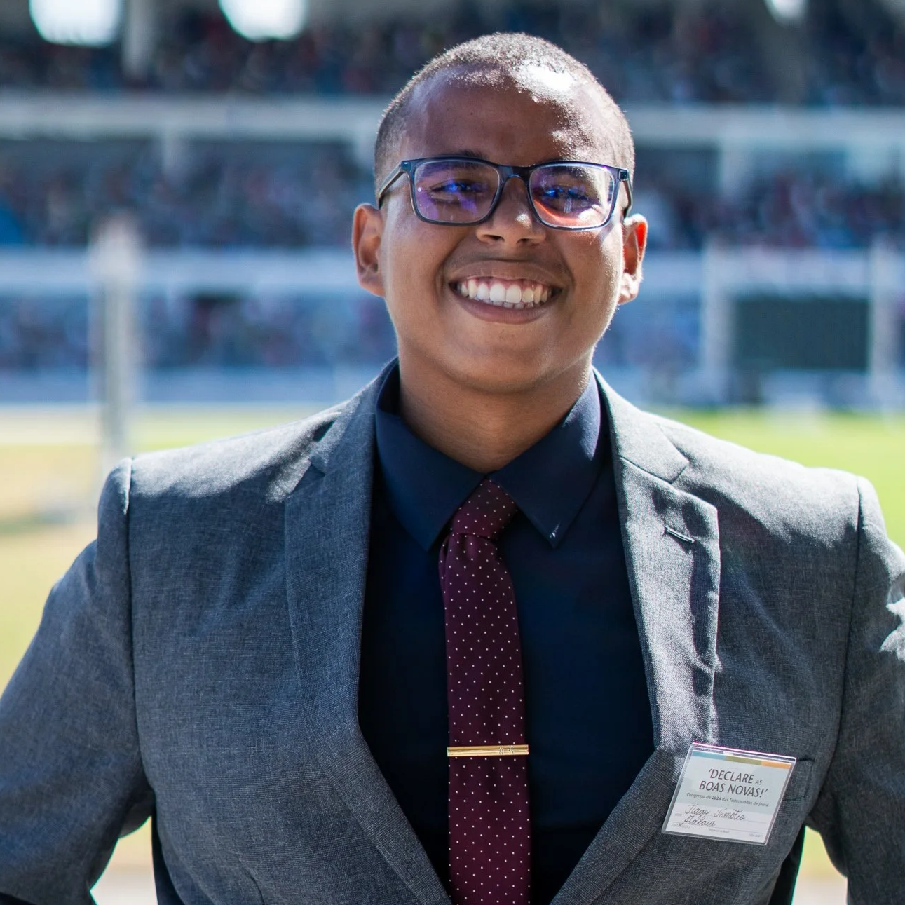 Homem sorrindo com braços cruzados, vestindo camiseta cinza escuro e uma pulseira inteligente, ao lado de uma parede branca com detalhes decorativos, ao ar livre com carros estacionados ao fundo.