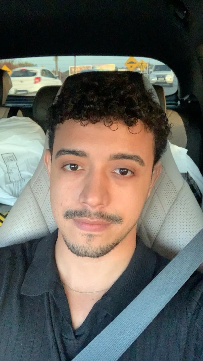 Homem sorridente de cabelo escuro, usando camisa branca, ao lado de uma janela ou uma porta de madeira, em um ambiente interno.