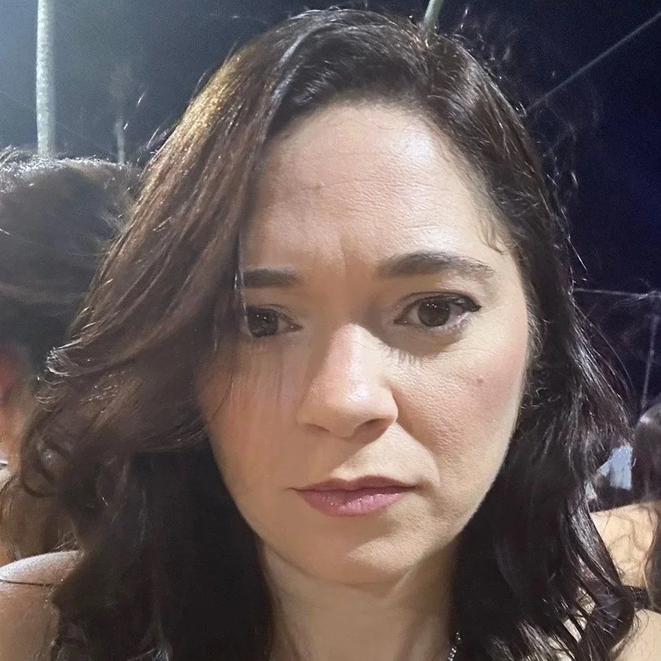 Mulher com cabelos longos e lisos, usando óculos de armação fina, sentada no interior de um carro, com fundo de janelas e assentos de carro visíveis.