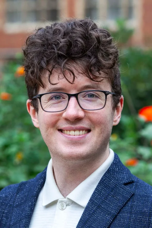 Portrait of a young man with curly brown hair, glasses, and a friendly smile, wearing a white shirt and a blue blazer, with a garden and building in the background.