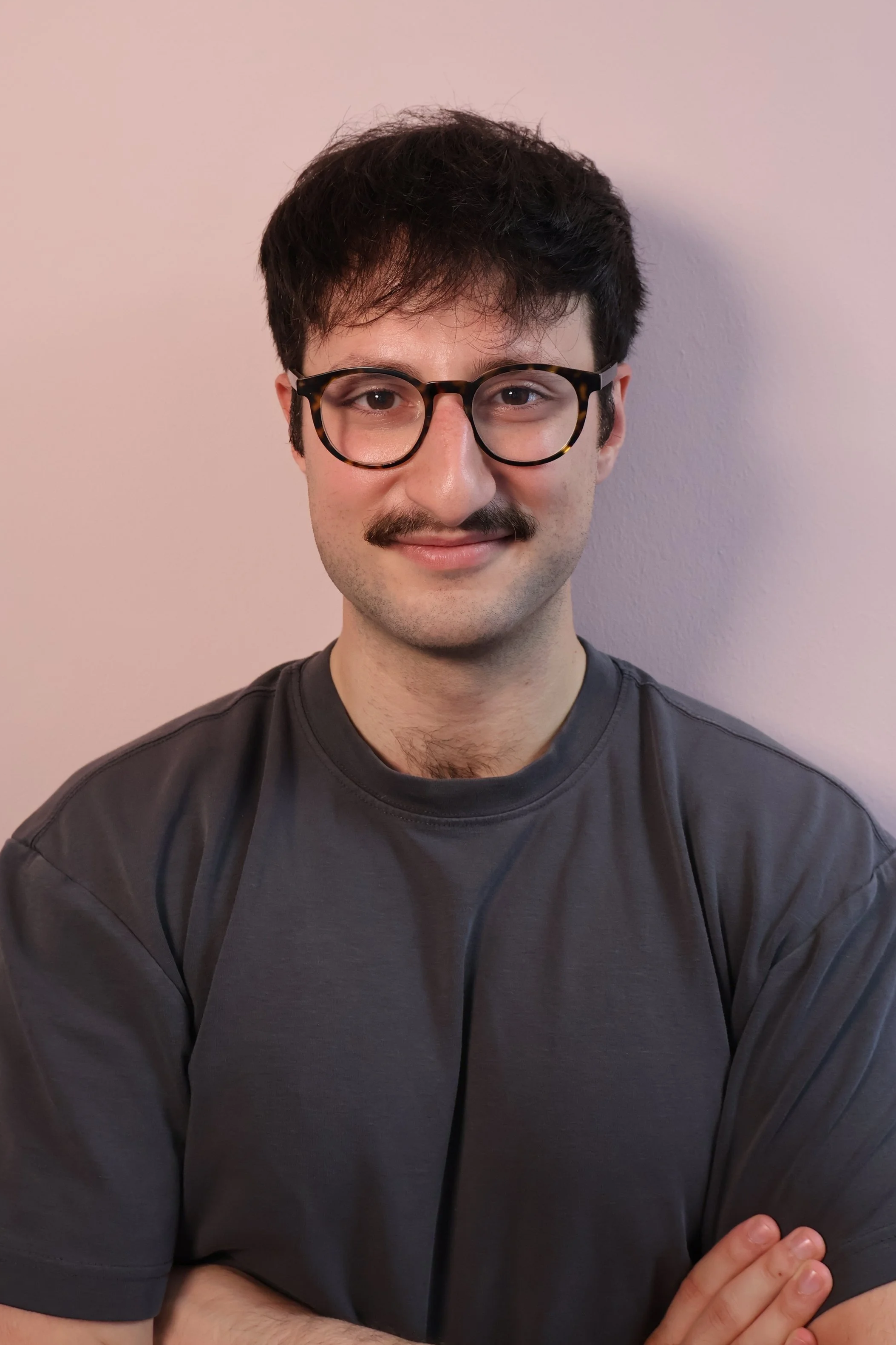 A young man with dark hair, glasses, and a mustache, wearing a dark gray T-shirt, standing against a plain light-colored wall.
