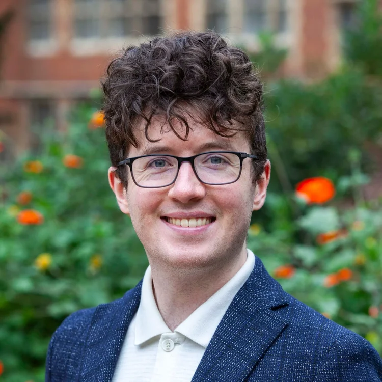A young man with curly brown hair and glasses, smiling outdoors with a background of green foliage and orange flowers.