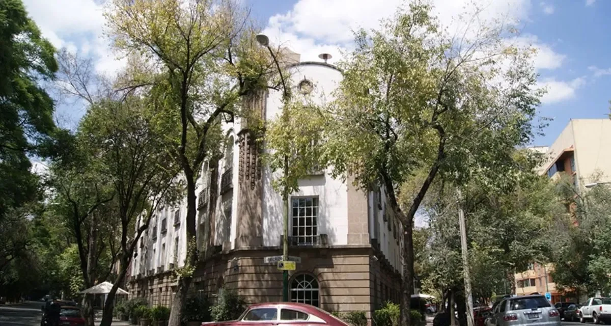 A street view of a historic building with white walls and a stone base, surrounded by tall trees with green leaves, parked cars, and a partly cloudy sky, in La Condesa Neighbourhood, Mexico City.