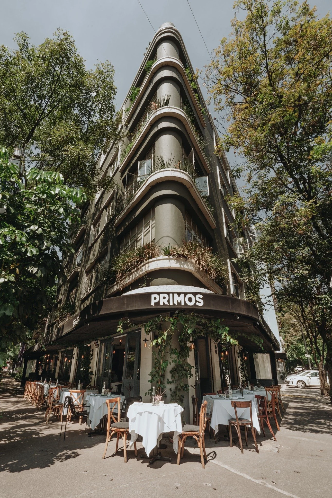 A multi-story building with a restaurant called Primos on the ground floor, outdoor dining tables with white tablecloths and wooden chairs, surrounded by trees and greenery, in La Condesa Neighbourhood, Mexico City.