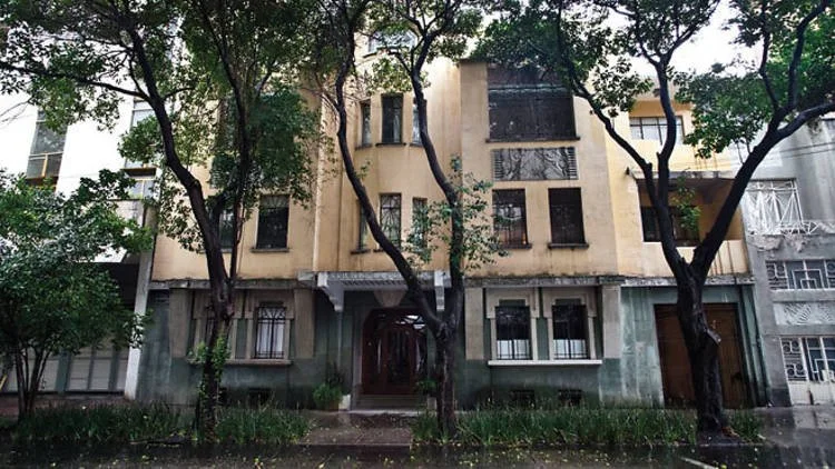 A multi-story residential building with a beige facade, black window frames, and a dark wooden front door, surrounded by trees and greenery on a rainy day, in La Condesa Neighbourhood, Mexico City.
