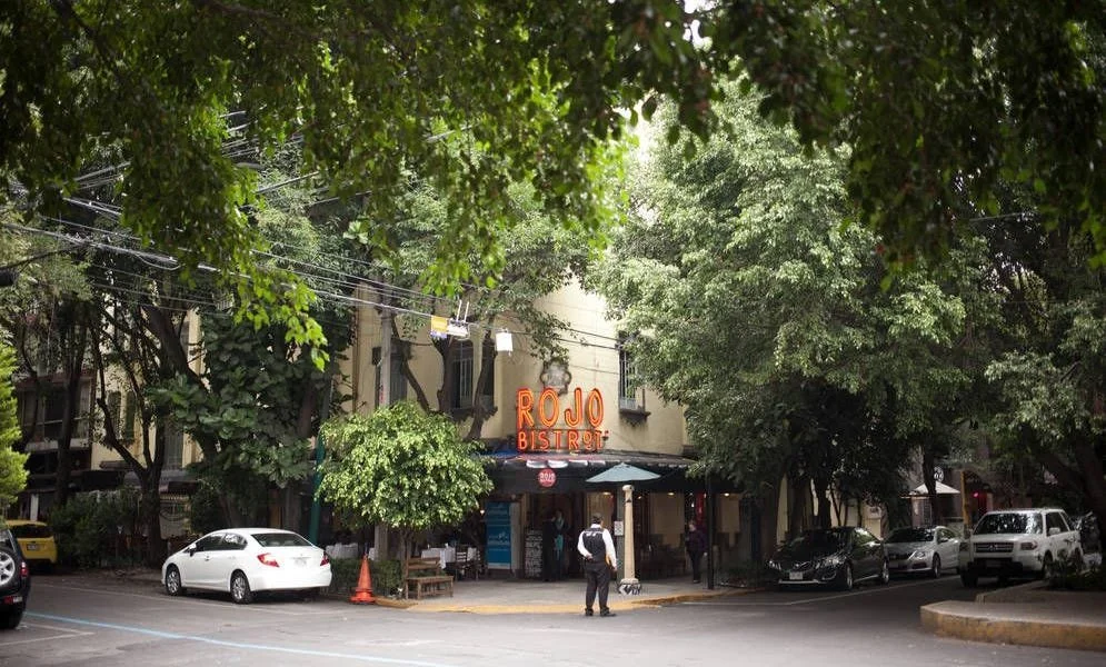 Street view of Rojo Bistro restaurant with trees, parked cars, and people walking, in an urban setting, in La Condesa Neighbourhood, Mexico City.