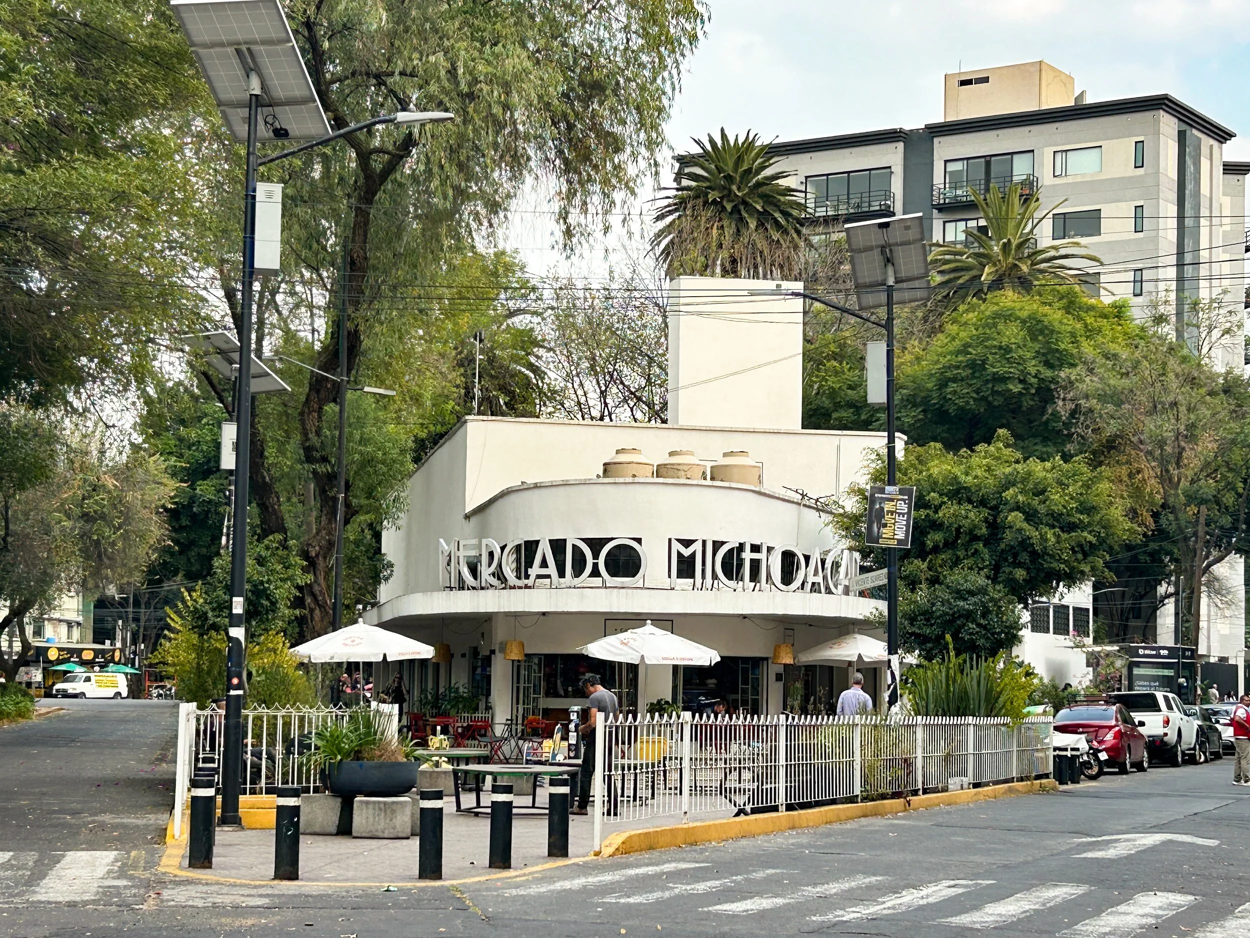 Mercado Michoacano restaurant with outdoor seating, umbrellas, and pedestrians on a busy street, surrounded by trees and modern buildings.