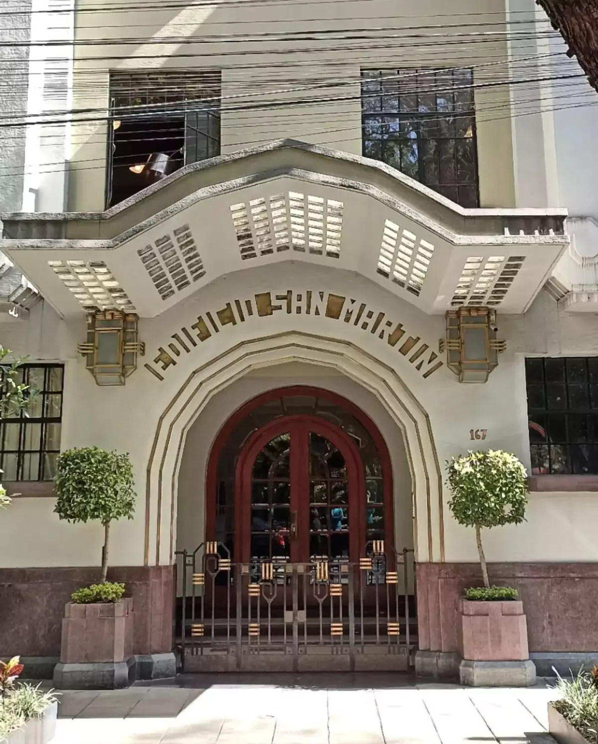 Building entrance with an arched doorway, red double doors, and two small trees in planters on either side, in La Condesa Neighbourhood, Mexico City.