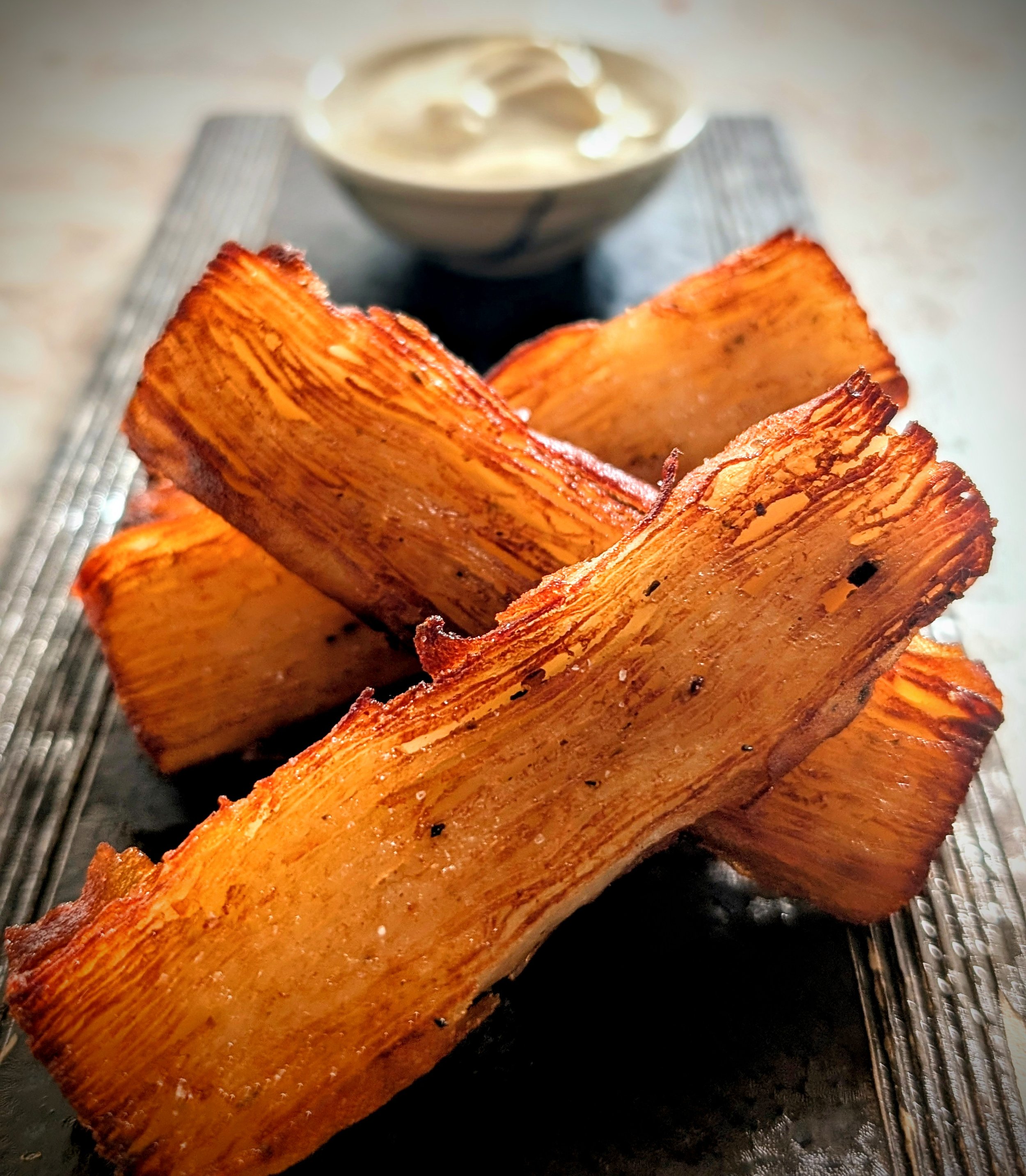 Close-up of crispy, golden-brown bacon strips on a grill with a small bowl of creamy sauce or dip in the background.