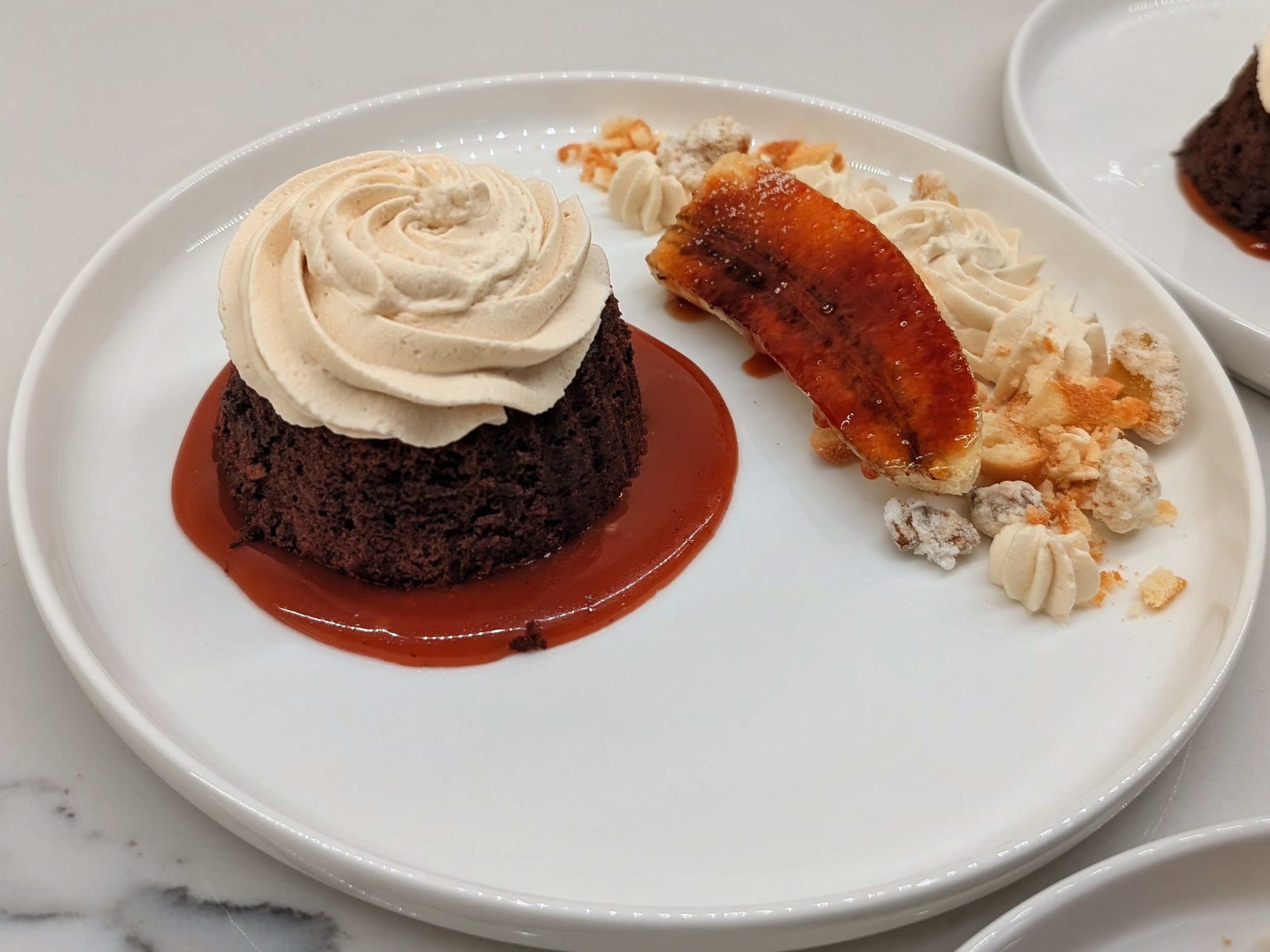 A white plate with a chocolate dessert topped with whipped cream and caramel sauce, alongside a slice of caramel flan, decorated with whipped cream and caramel, and irregular crumbs or cookies, on a marble surface.