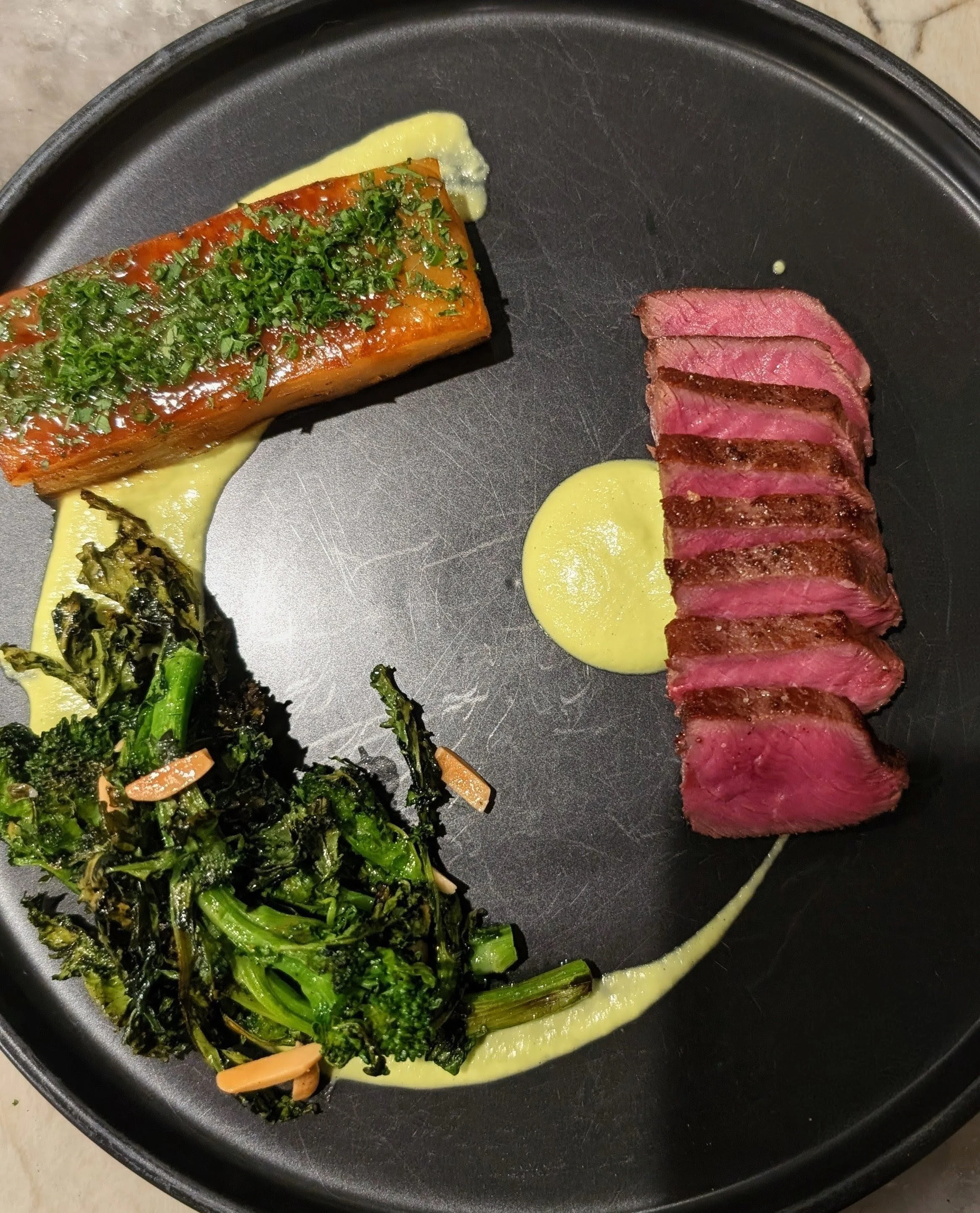 A black plate holds a row of sliced medium-rare steak, a piece of salmon topped with green herbs, and a serving of cooked kale with sliced almonds, all drizzled with a light yellow sauce.