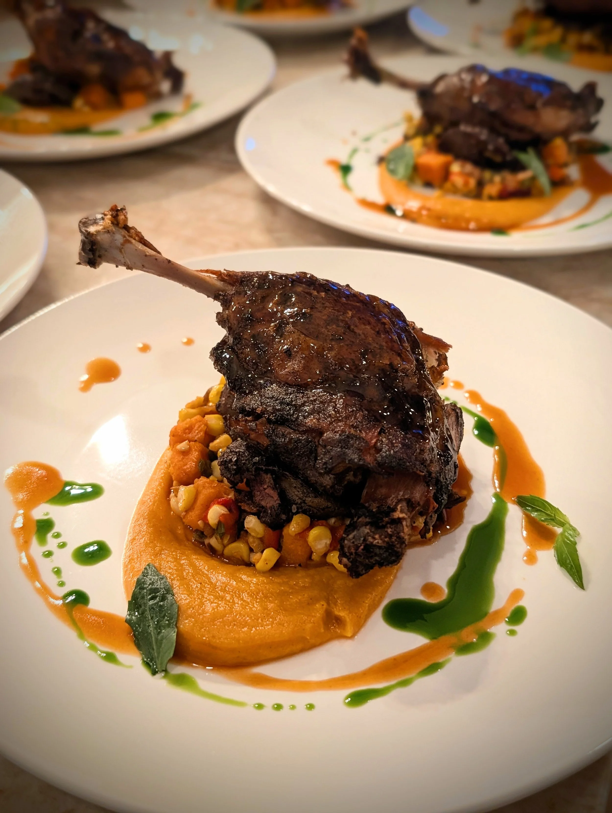 Close-up of a gourmet dish featuring a braised meat (likely beef or lamb) served over a bed of pureed vegetables, garnished with colorful sauces and herbs.