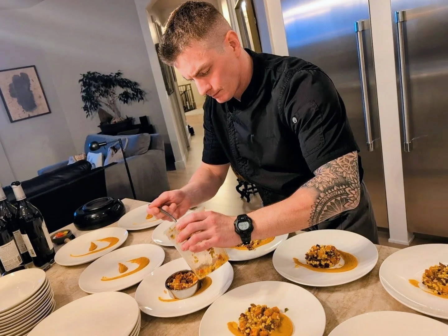A male chef with tattoos on his left arm, wearing a black uniform and watch, is garnishing plates of food in a modern kitchen, with several white plates containing a dish with orange sauce and a mixture of ingredients.