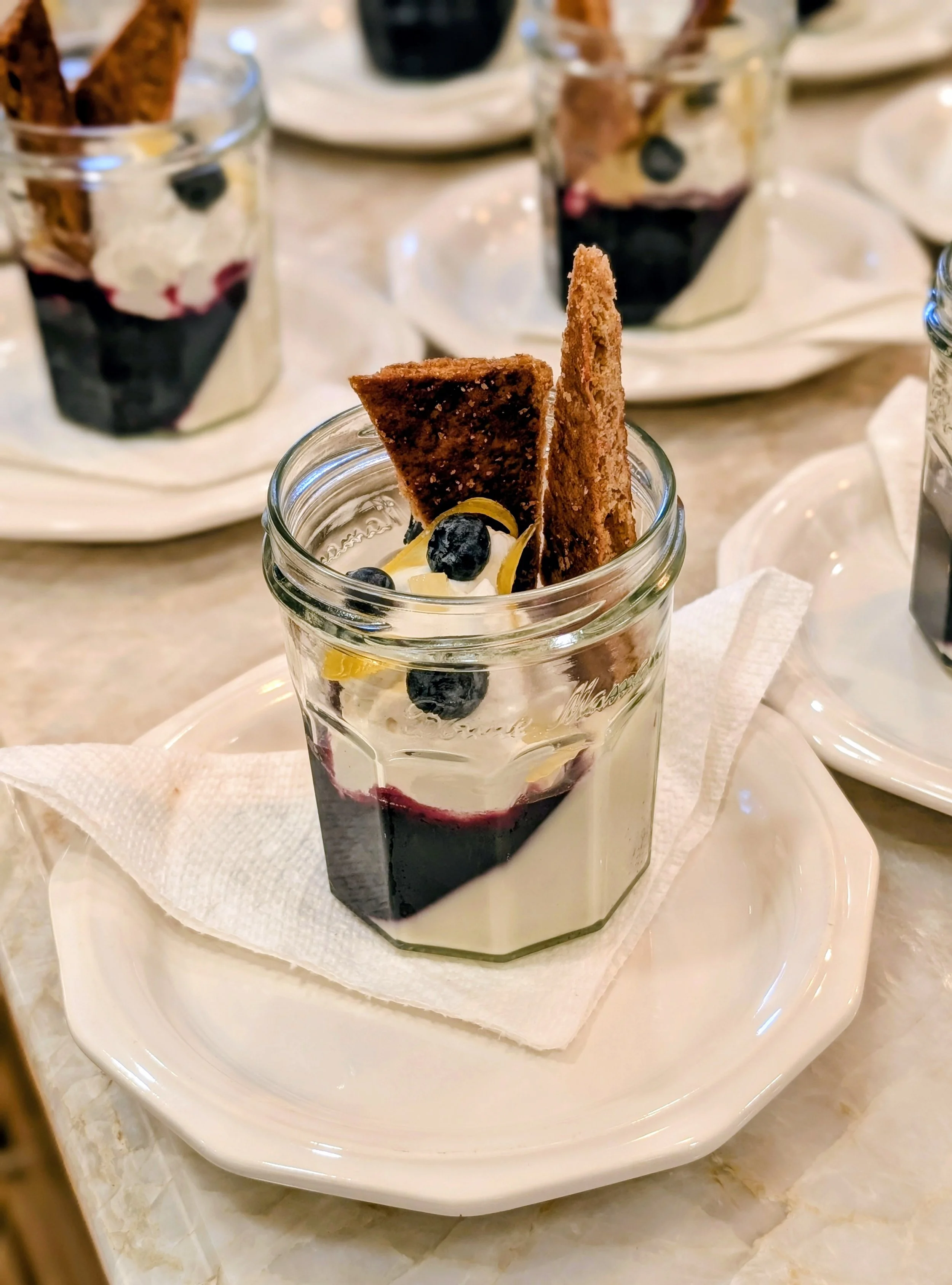 Glass jar filled with layered dessert including blueberries, whipped cream, and biscuit cookies, placed on a white plate with napkins.
