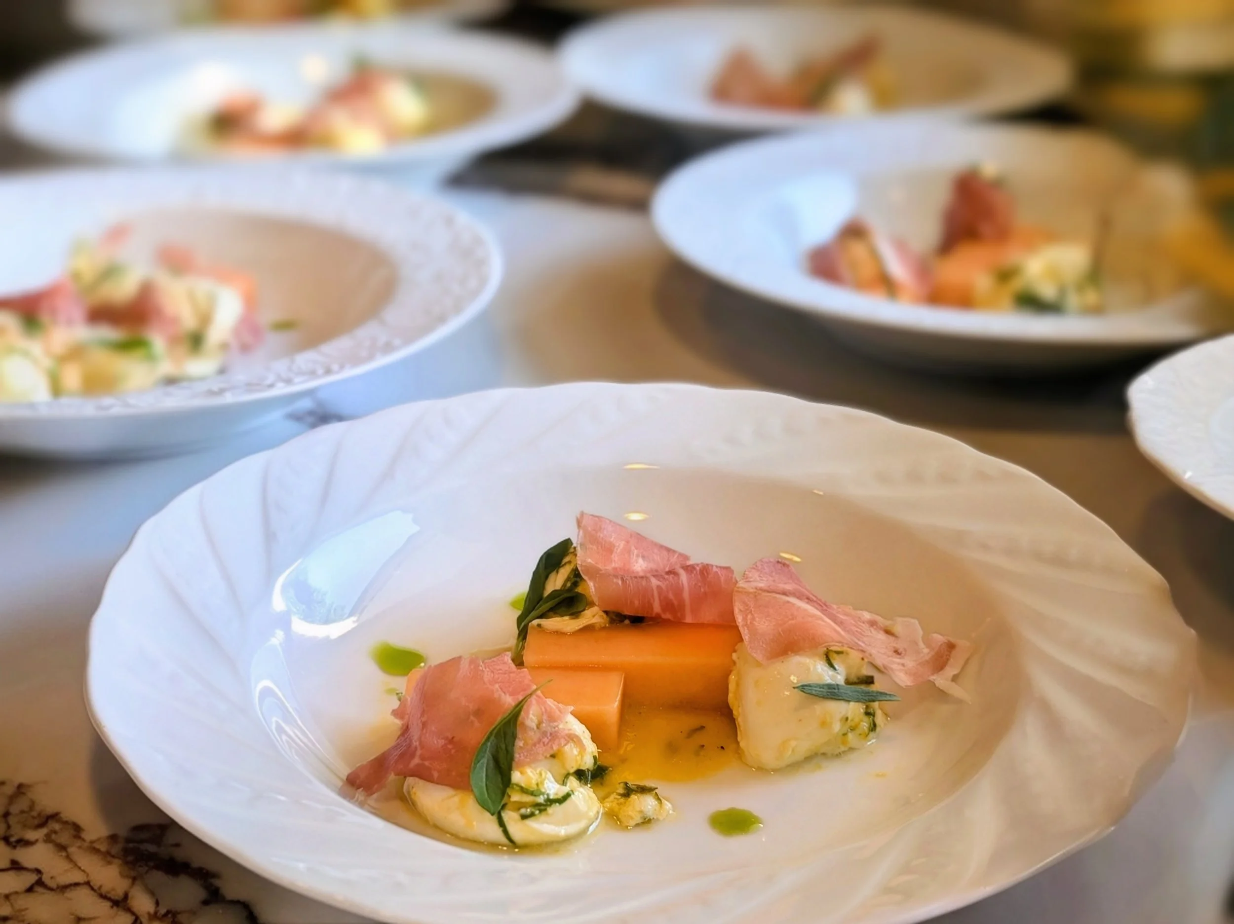 A white plate of gourmet appetizers with slices of prosciutto, cheese, and herbs, garnished with green oil or herb oil drizzle.