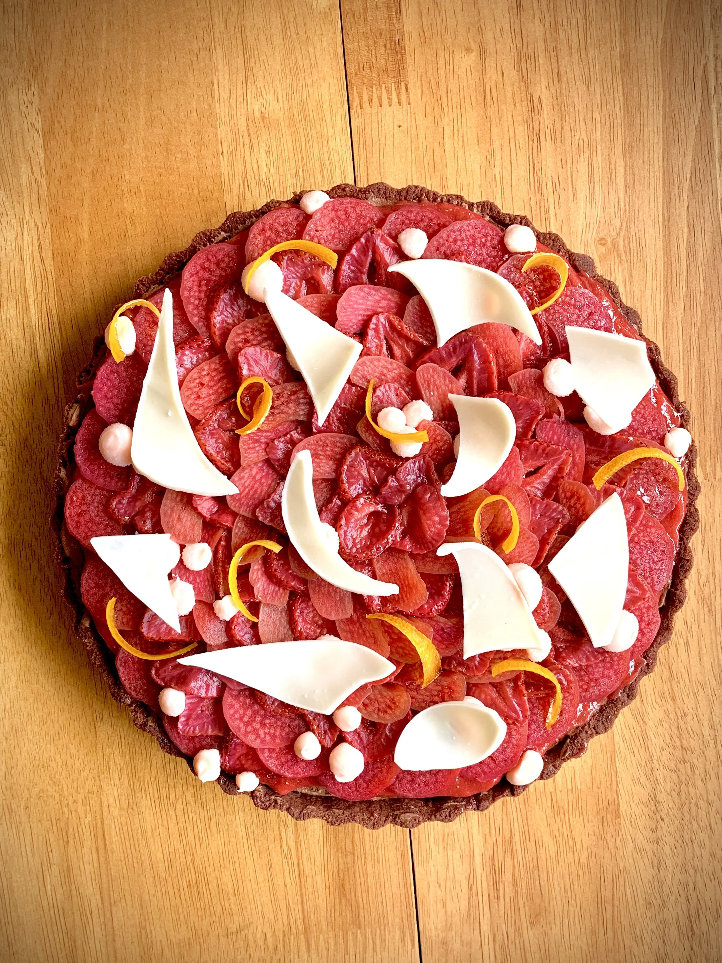 Round tart with a chocolate crust, topped with sliced red fruits, white chocolate pieces, and small white and yellow decorations on a wooden surface.