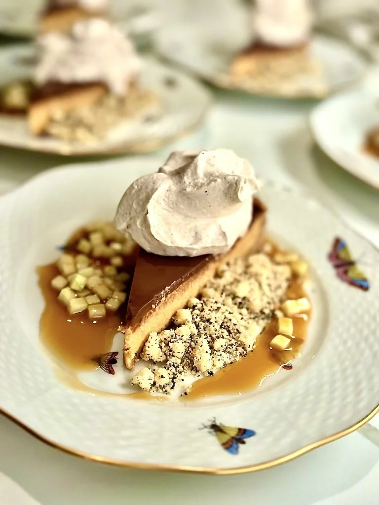 A slice of chocolate and graham cracker cheesecake topped with whipped cream on a decorative plate with honey, chopped nuts, and crumbled graham cracker crumbs, with more slices of cheesecake blurred in the background.