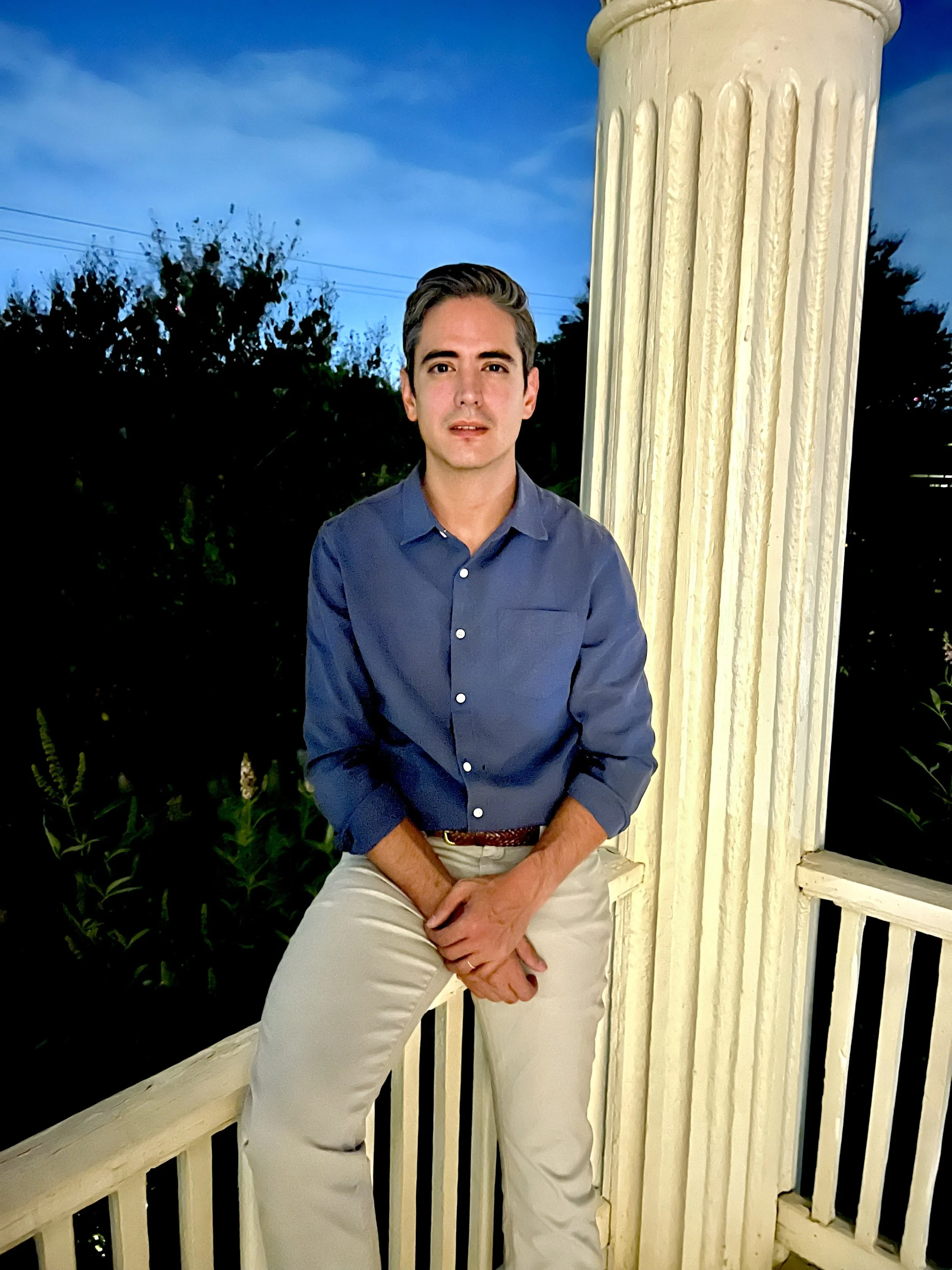 A young man with dark hair, wearing a blue button-down shirt and light-colored pants, is sitting and leaning against a white porch railing with a large, white, fluted column next to him. The background includes trees and a blue sky with some clouds, suggesting it is evening or dusk.