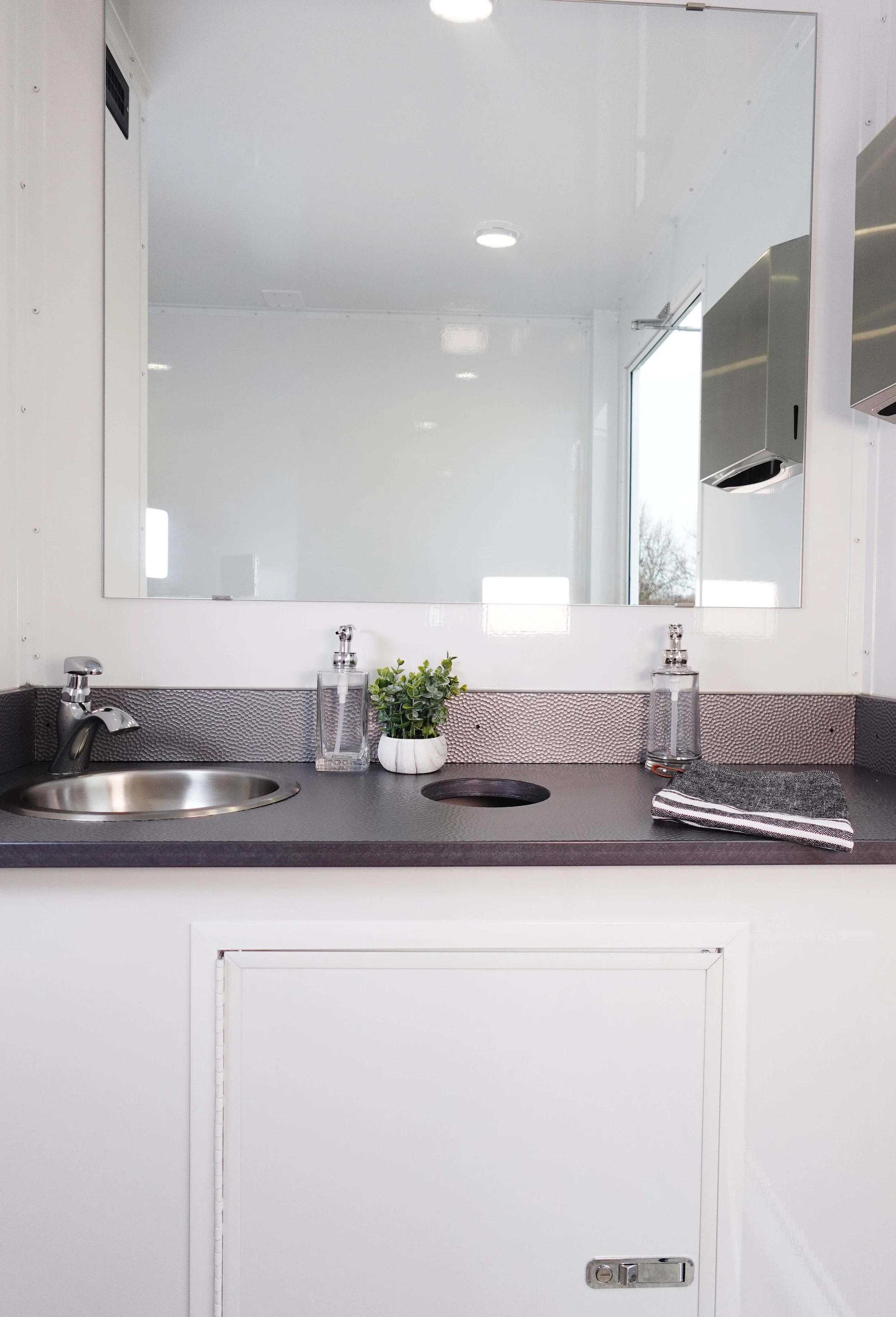 A clean, minimalist sink area with a small round stainless steel sink, a soap dispenser, a striped cloth, and a potted plant on a dark textured countertop. A large mirror reflects a bright white space with windows and ceiling lights.