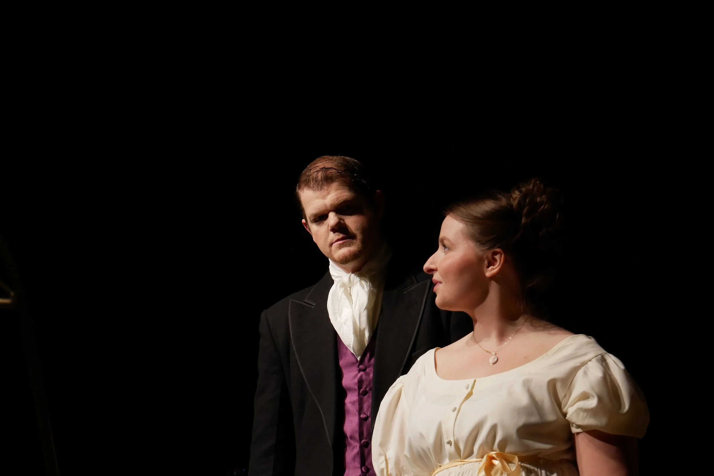 Two actors in period costumes perform a scene on stage with a dark background, one man with serious expression and woman with a slight smile