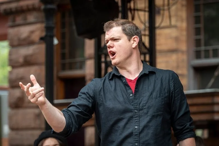 A man with short light brown hair is speaking passionately, gesturing with his right hand, while standing outdoors in front of a wooden building.