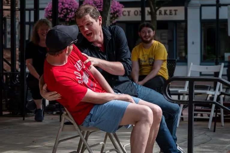 A man laughing and holding a seated boy wearing a red shirt and cap, with two women smiling and sitting outdoors nearby.