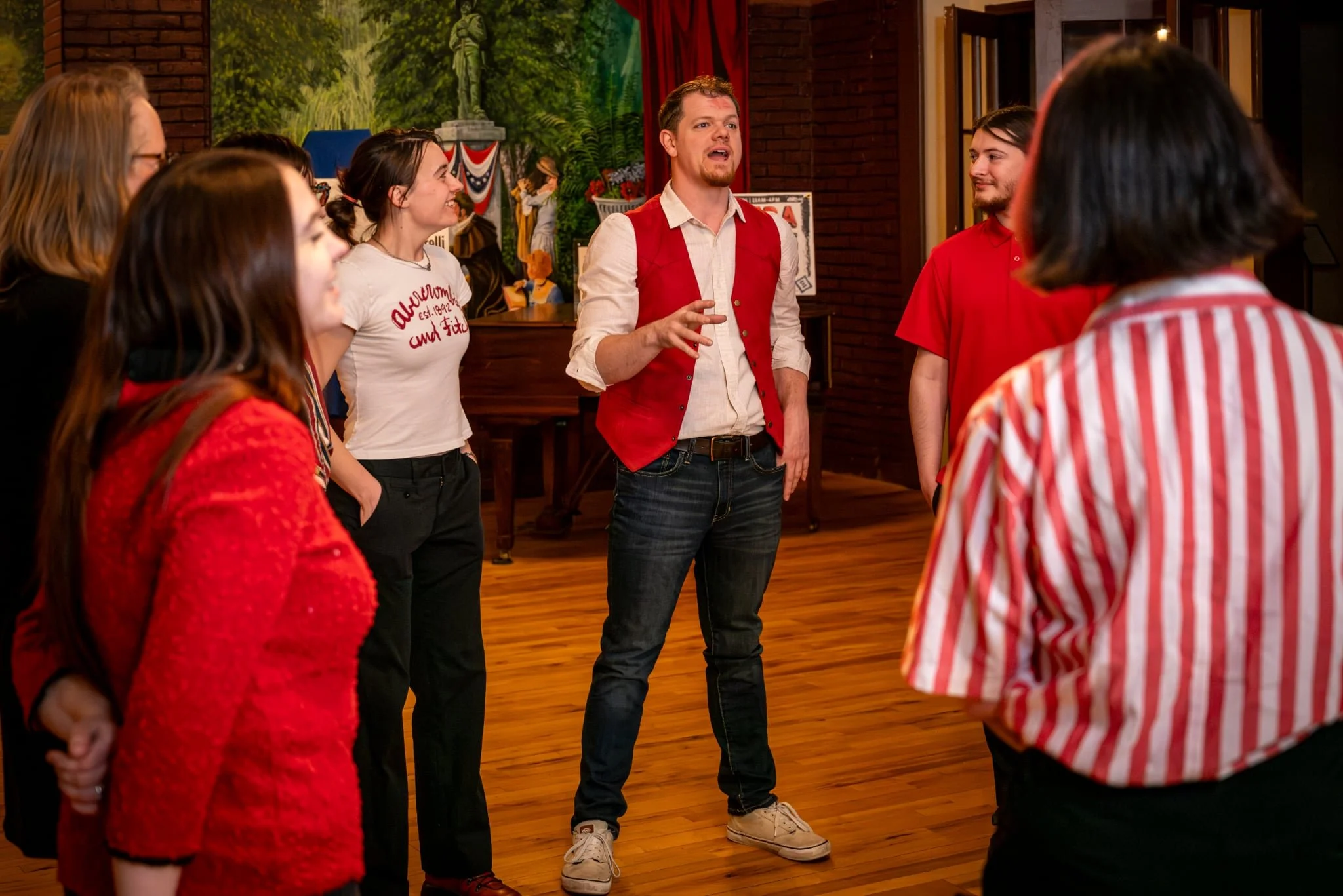 A man in a red vest and white shirt speaking to a group of young people indoors.