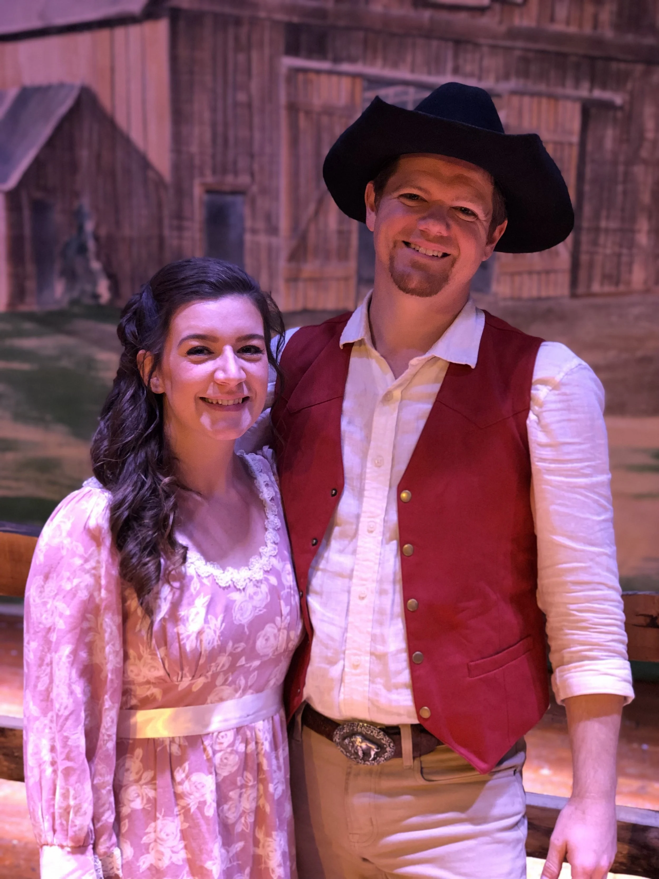 A man and woman dressed in western-style costumes, smiling and standing together in front of a barn backdrop.