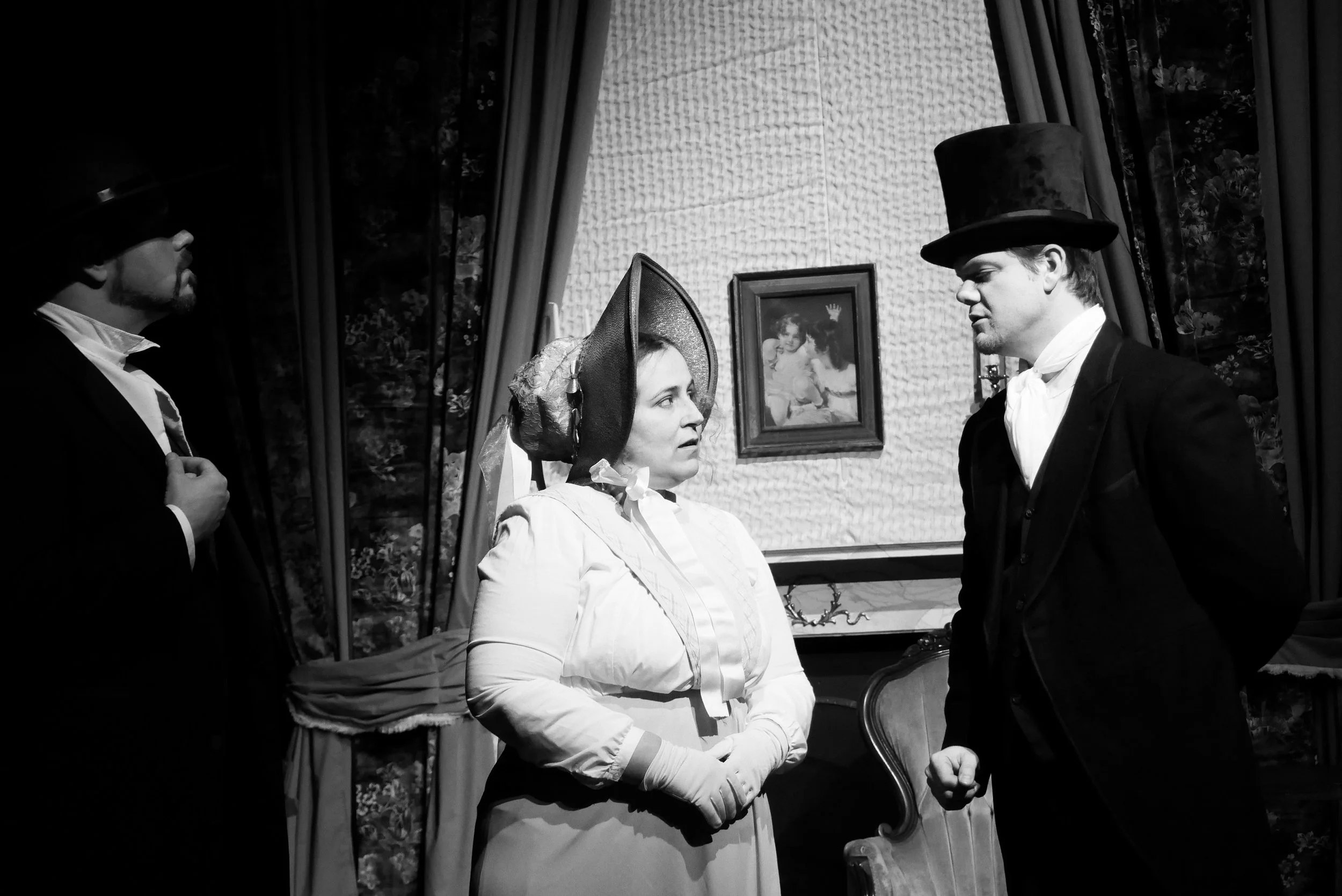 Three people dressed in 19th-century clothing engaging in a conversation in a Victorian-style room with curtains, a framed portrait on the wall, and antique furniture.