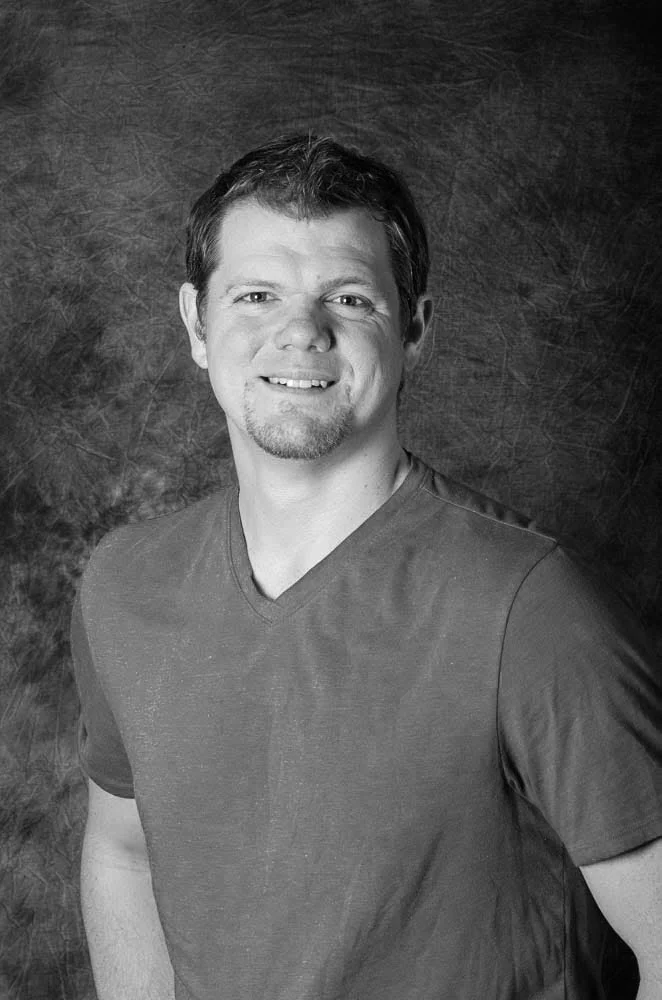 Black and white portrait of a smiling man with short hair and a beard, wearing a casual T-shirt, standing against a textured dark background.