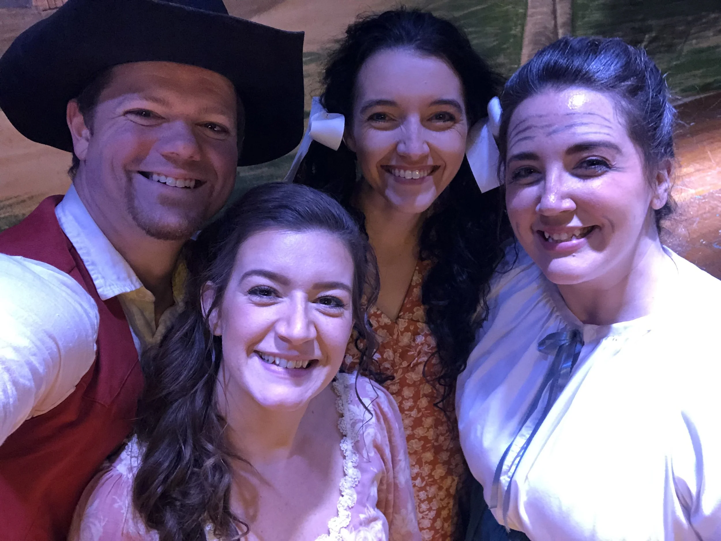 Four people dressed in costumes, smiling for a group selfie, with a man wearing a large black hat and a woman with a white bow in her hair.