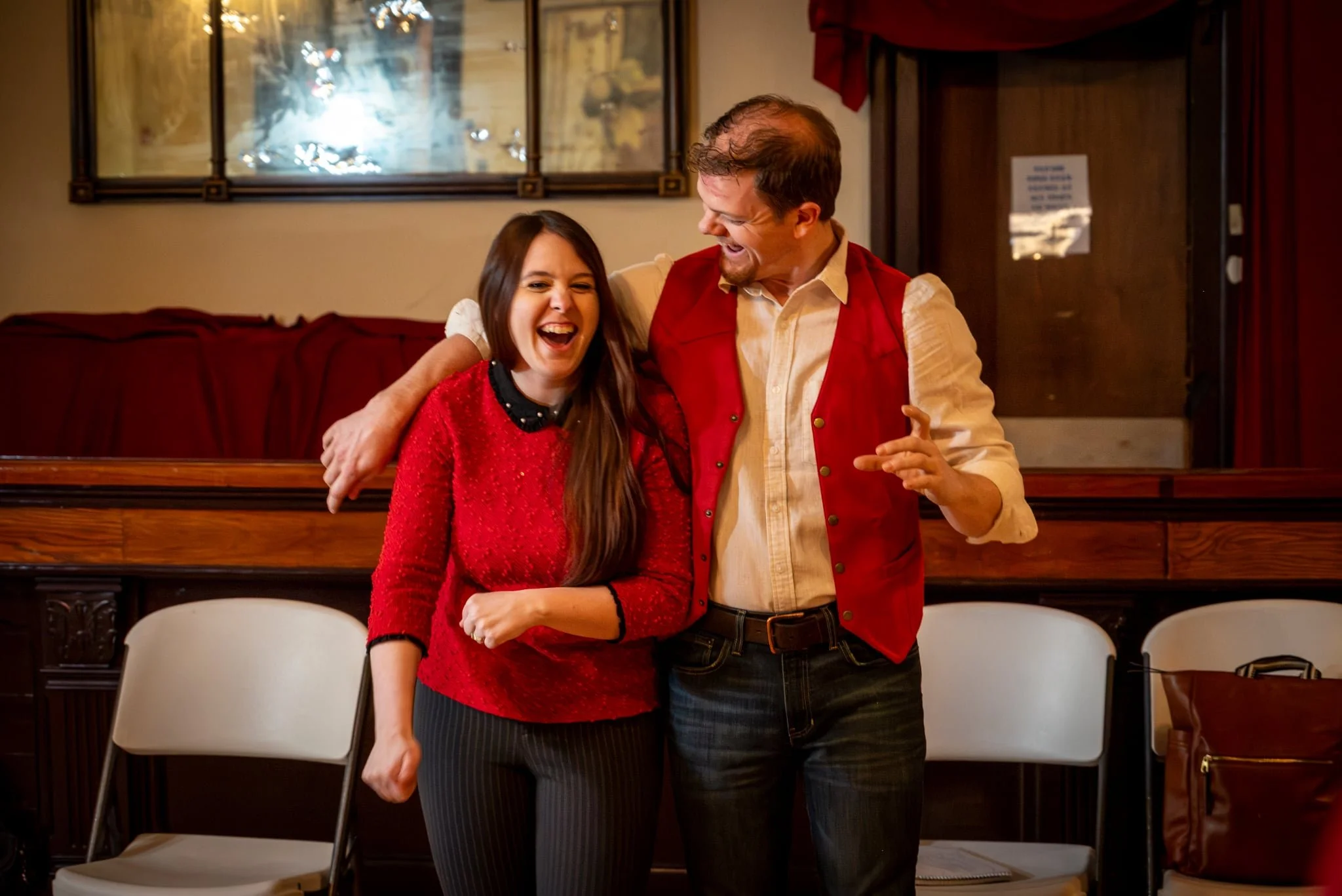 A man and a woman are laughing and enjoying each other's company indoors. The woman has long brown hair and is wearing a red sweater and black pants. The man has short brown hair and is wearing a cream shirt with a red vest. He has his arm around the