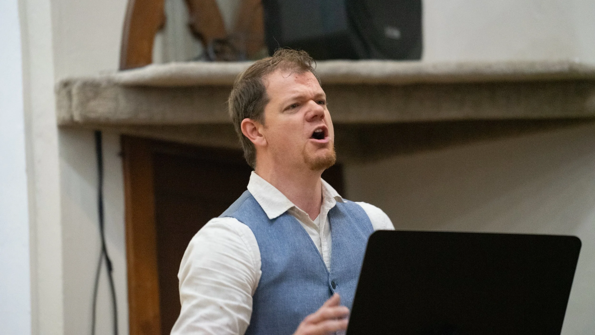 A man with short brown hair, wearing a light shirt and a blue vest, appears to be singing or speaking passionately while standing behind a laptop.