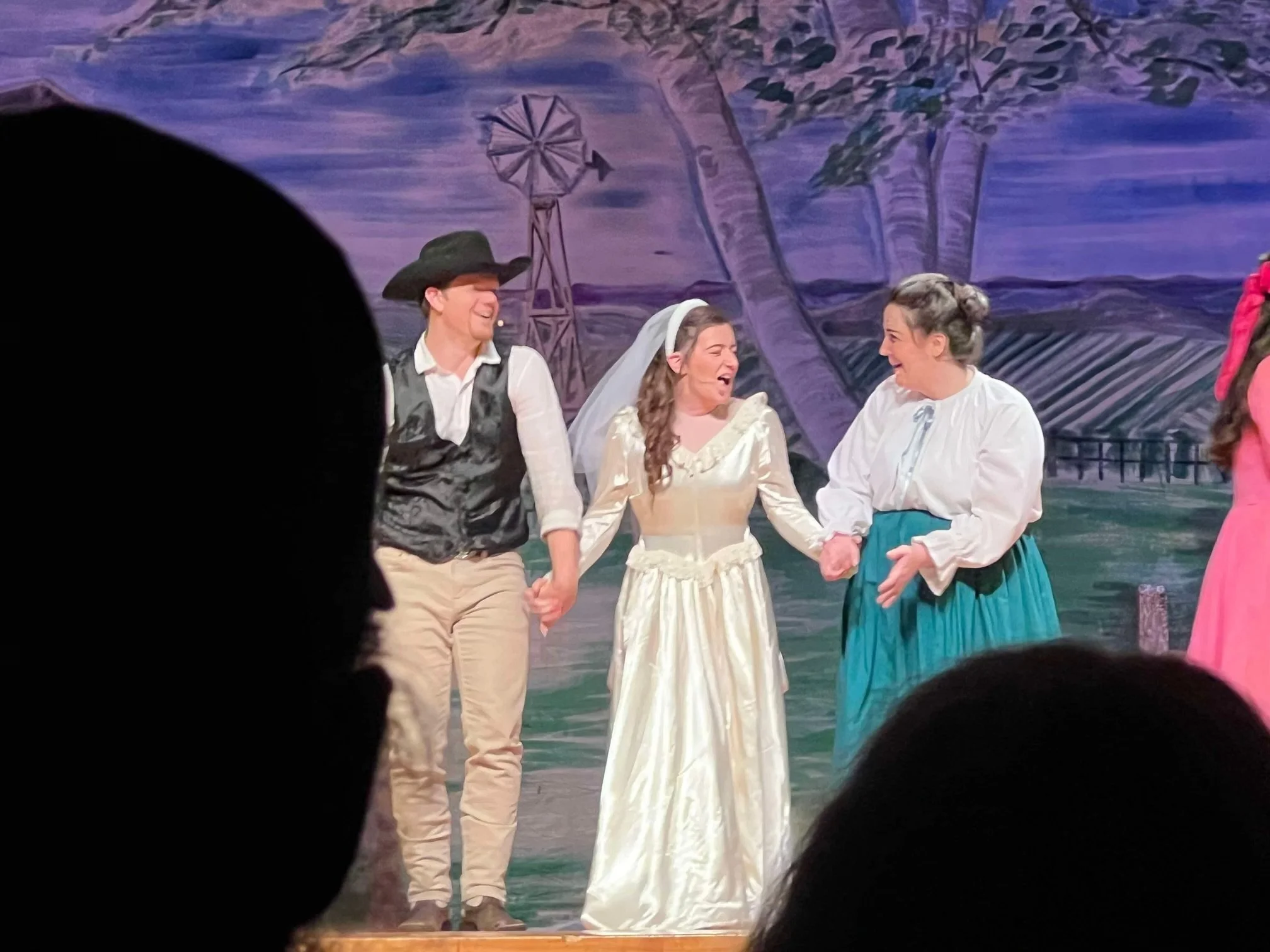 Four actors in period costume performing on stage outdoors with a painted rural background, holding hands and smiling.