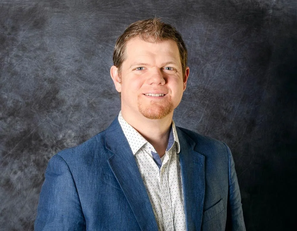 A man with short light brown hair, a goatee, wearing a blue blazer and a white shirt with small dark polka dots, standing against a dark textured background.