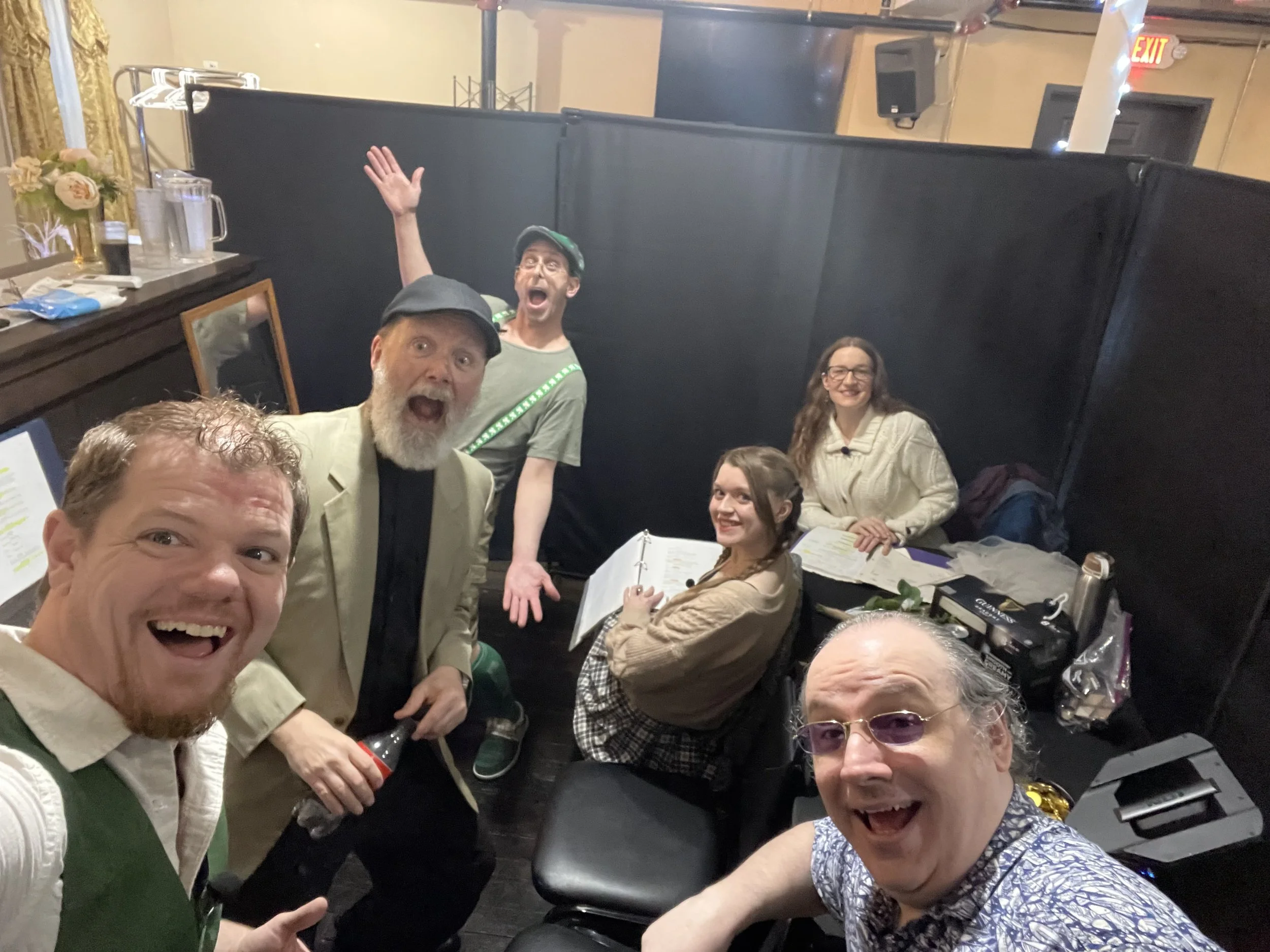 Group of six people smiling and enjoying themselves at a social gathering, some sitting and some standing, with a black partition and a bar area in the background.