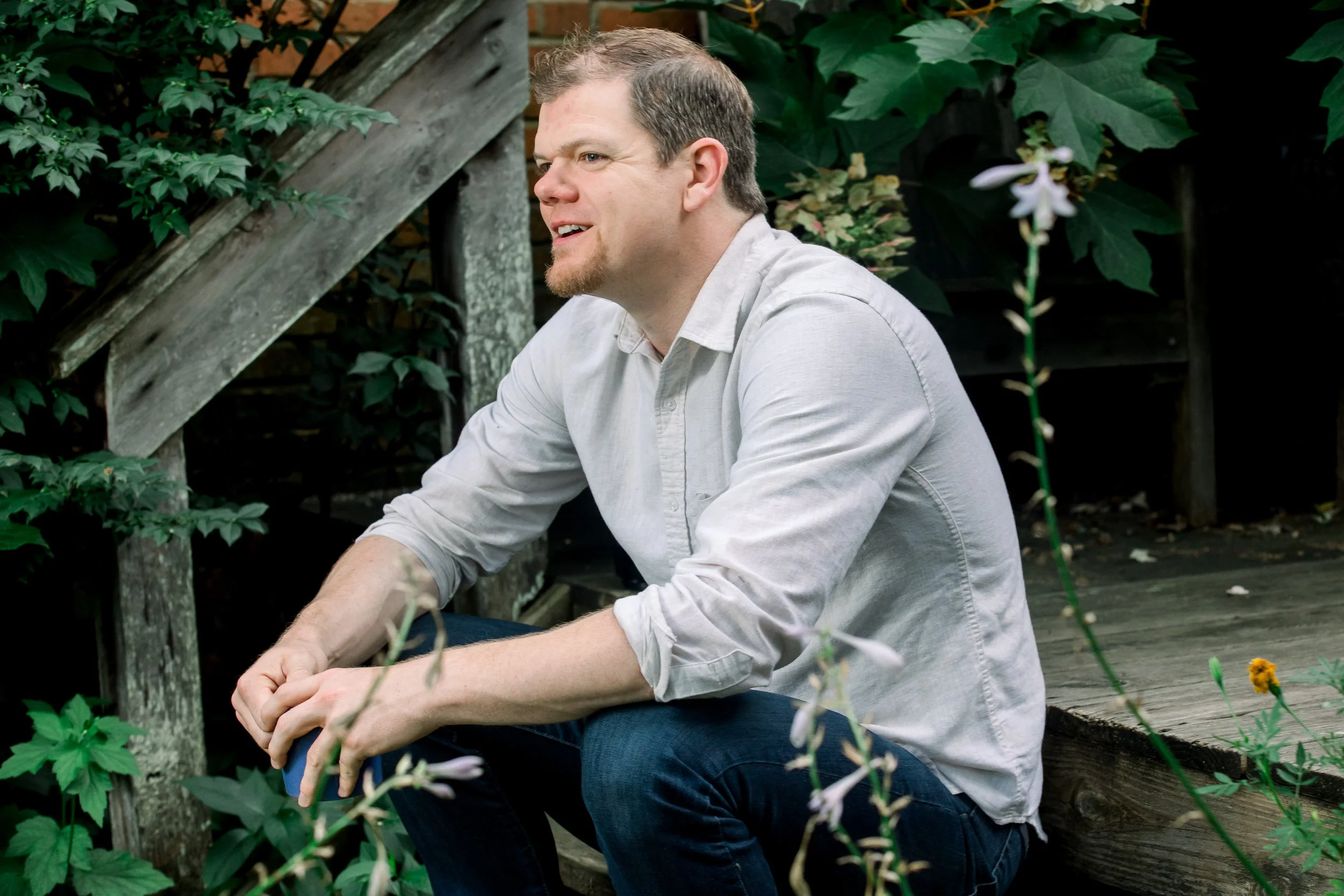 A man with light brown hair and a beard, wearing a light gray shirt and dark jeans, sits on a wooden platform in a garden surrounded by green plants and foliage.