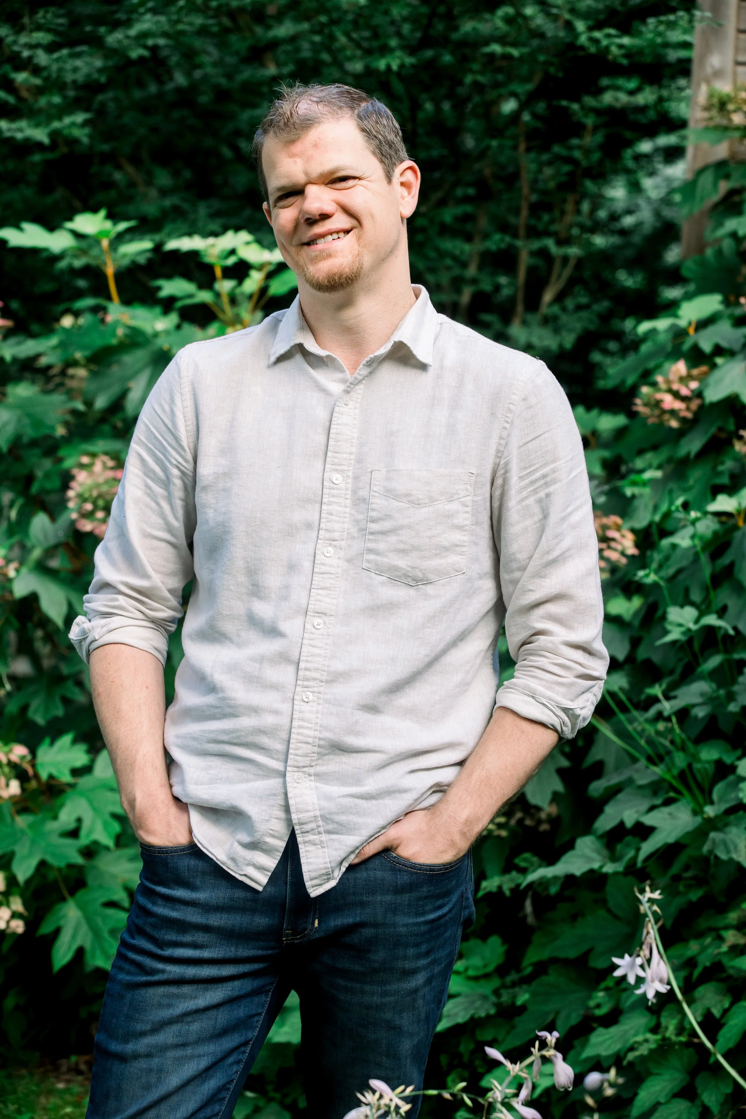 A man standing outdoors in front of green foliage, smiling with hands in pockets, wearing a light gray button-up shirt and dark jeans.