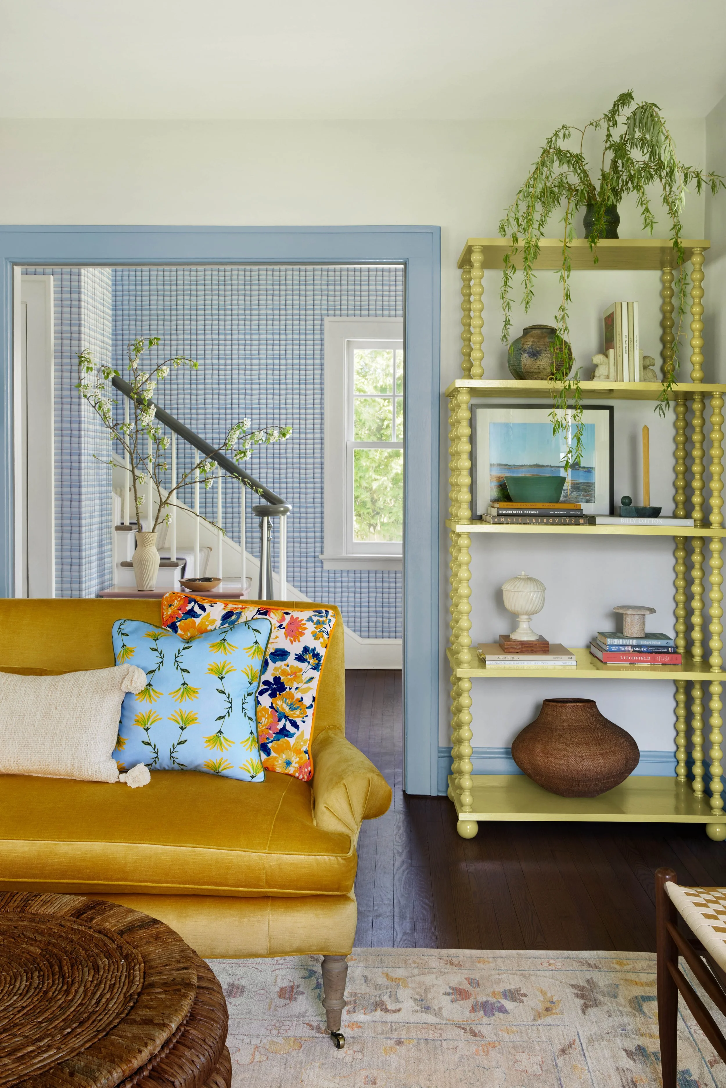 A colorful living room with a mustard yellow vintage sofa, decorative pillows, a rounded wicker coffee table, a light yellow bookshelf with books and decorative items, and a window with greenery outside. The walls feature blue and white checkered wallpaper.