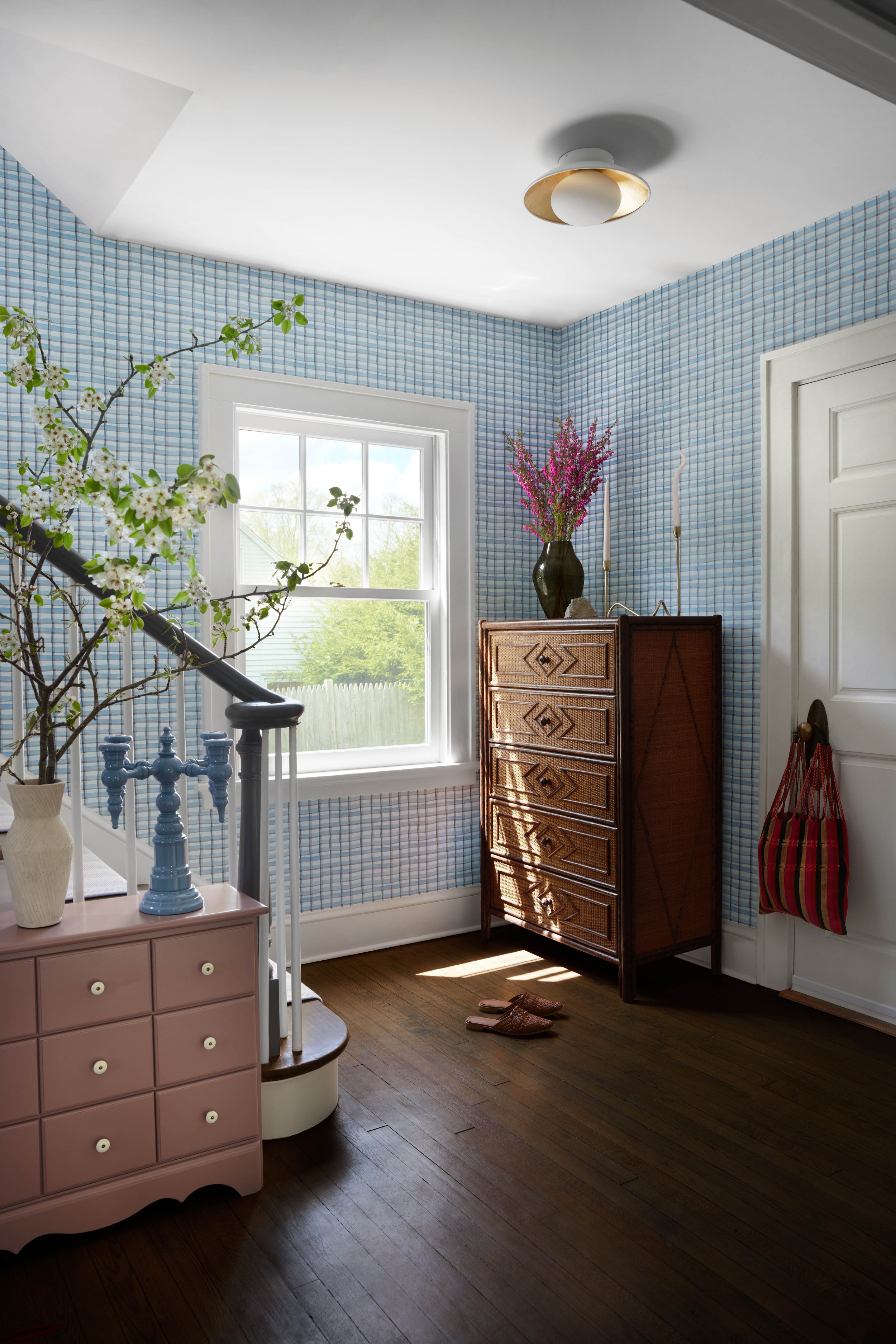 Interior of a room with a pink dresser, a wooden chest of drawers, a window with outdoor trees visible, and a pair of slippers on the wooden floor.